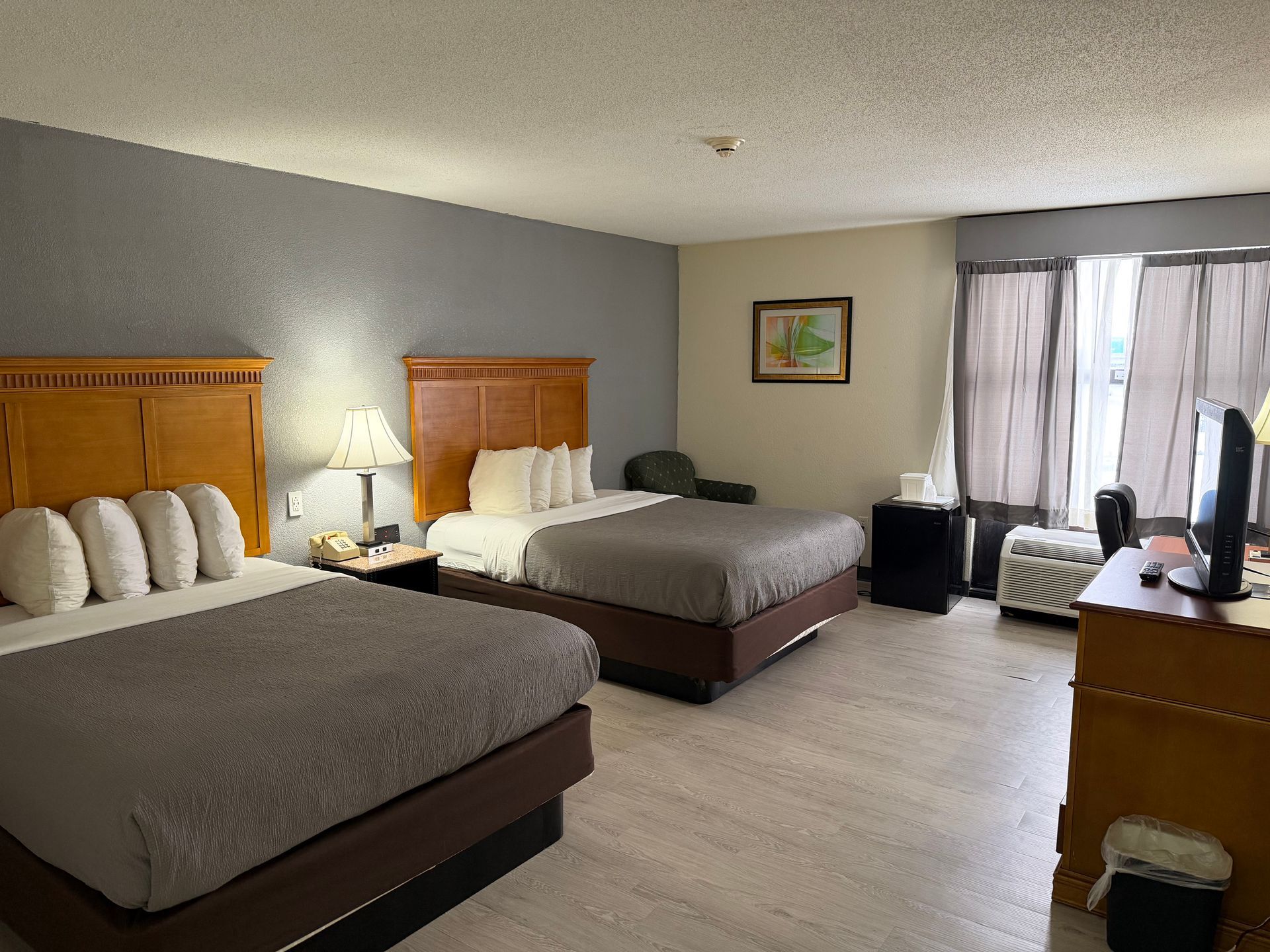 Hotel room with two beds, nightstands, a chair, and a desk near a window. Beige and gray color scheme.