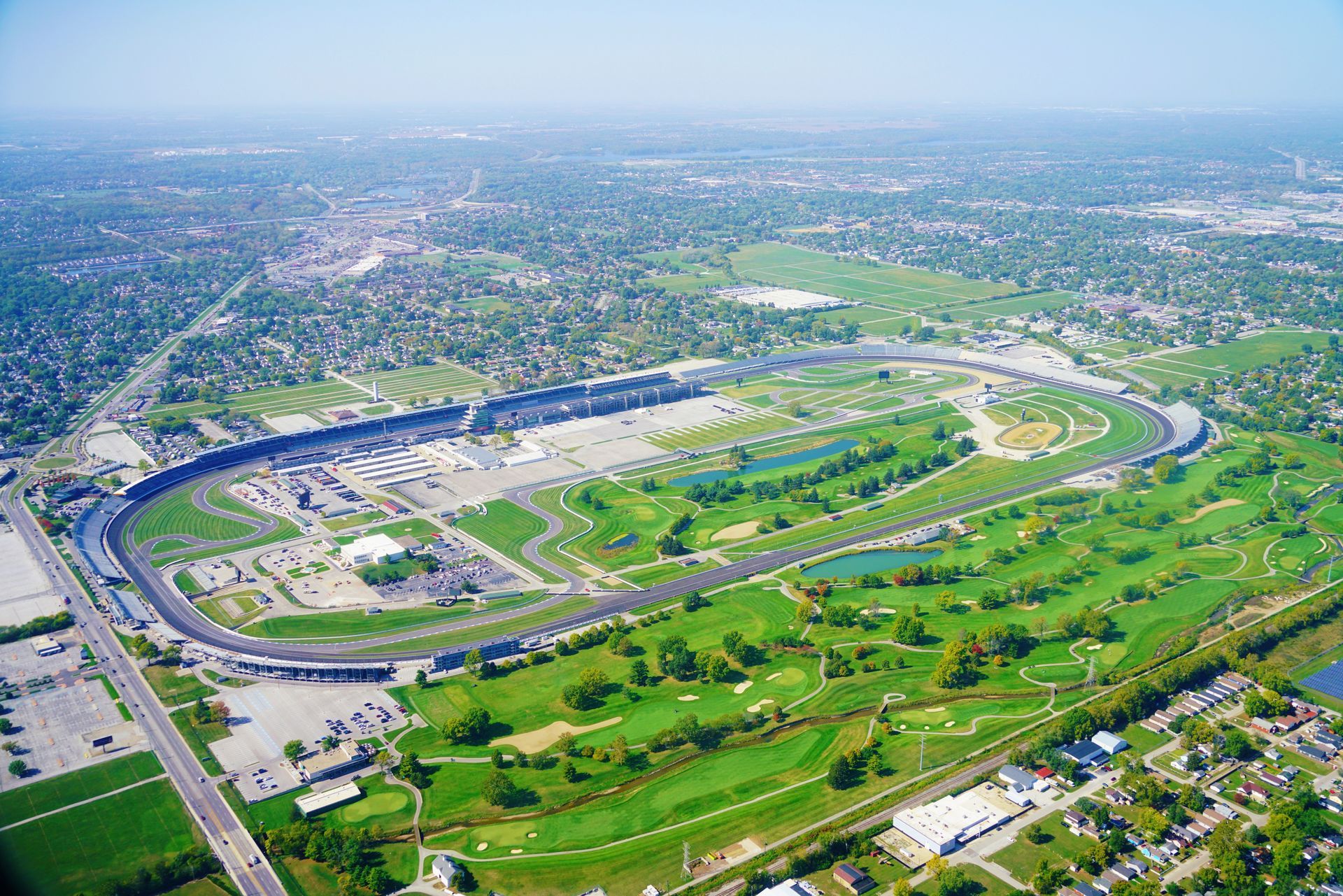 Aerial view of the Indianapolis Motor Speedway, a large oval race track with surrounding green spaces and buildings.