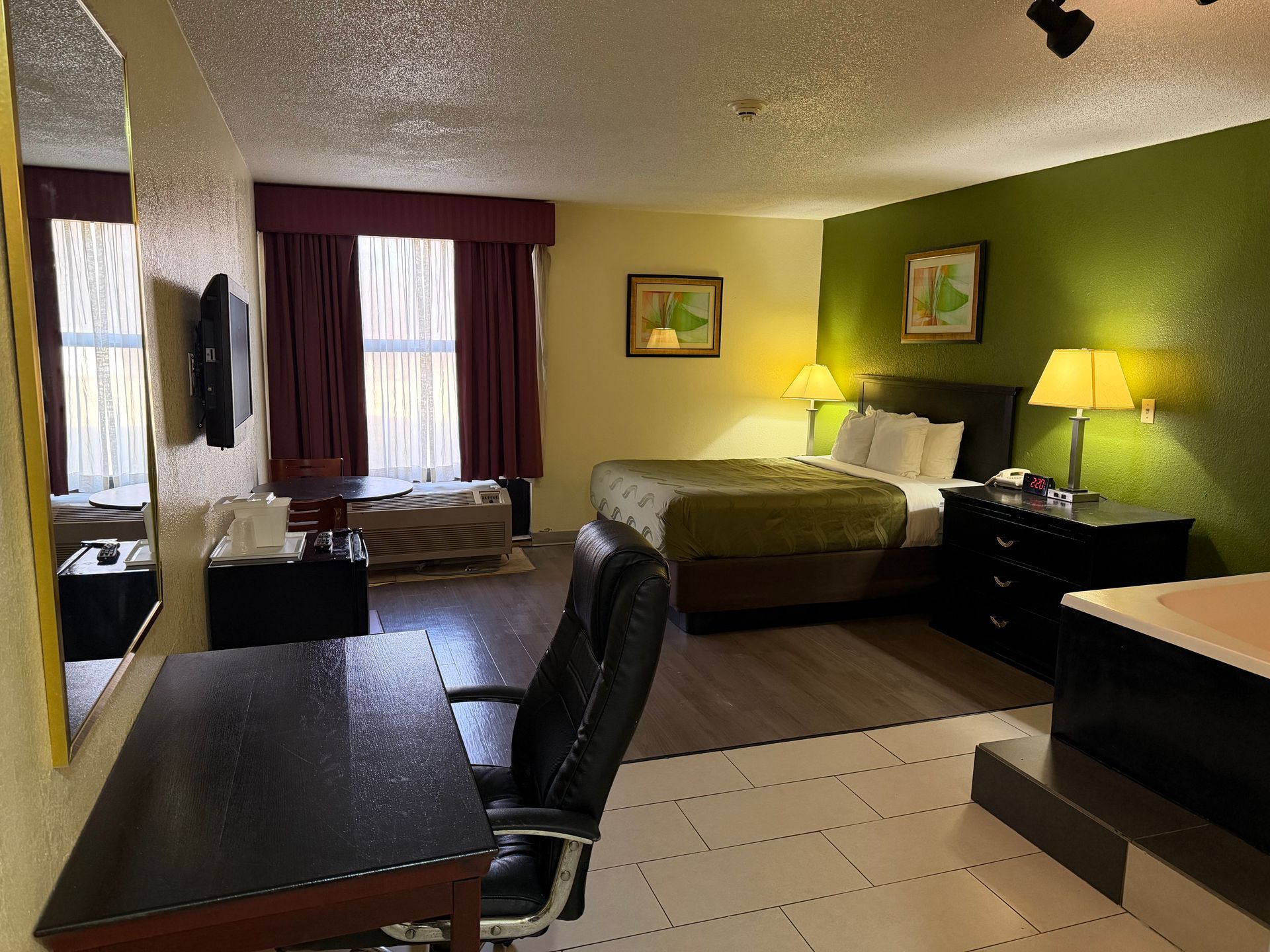 Hotel room with a bed, desk, TV, and window. Green and beige walls with dark furniture.