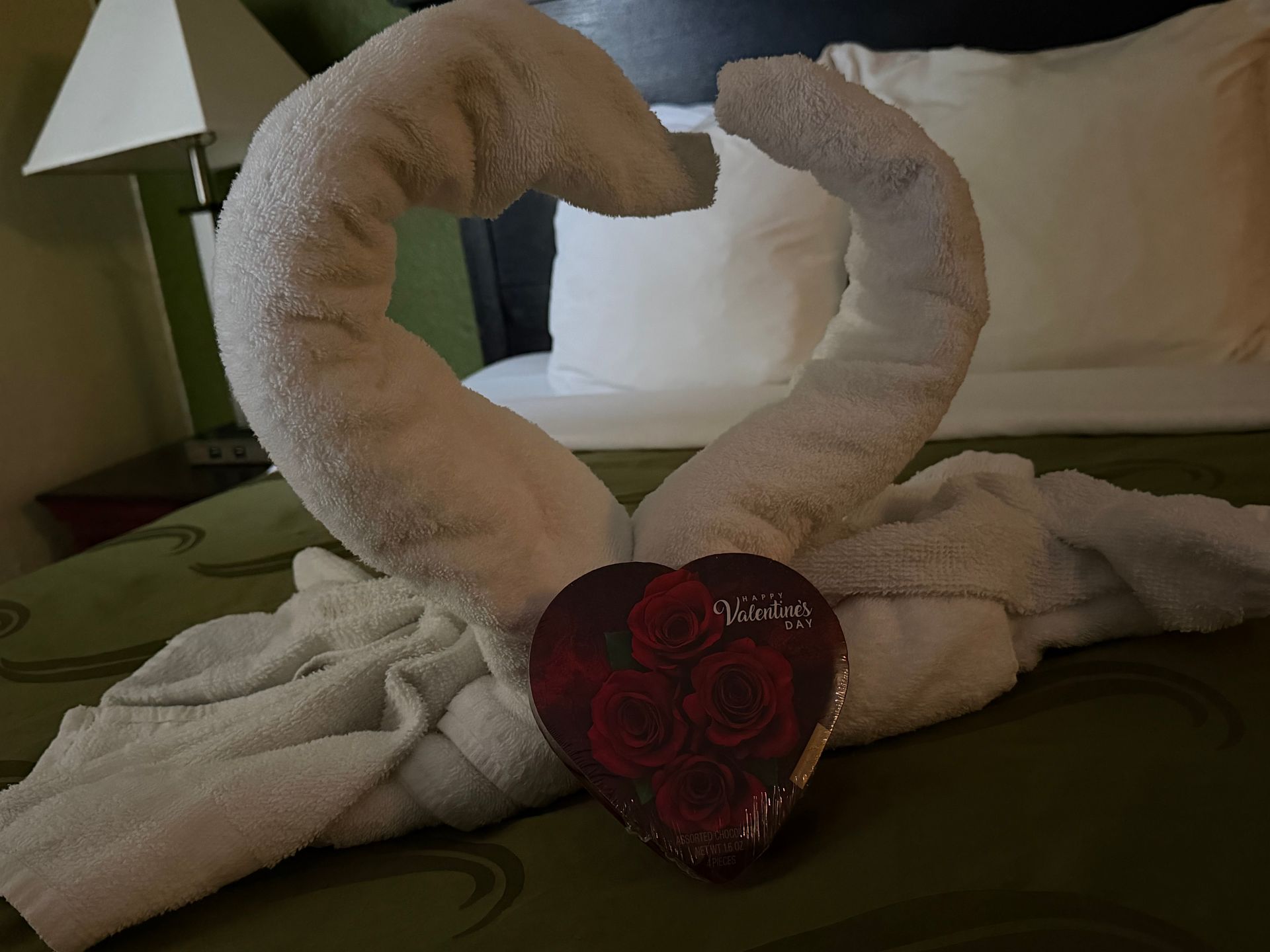 Towel swan with heart-shaped candy box on bed, hotel room setting.
