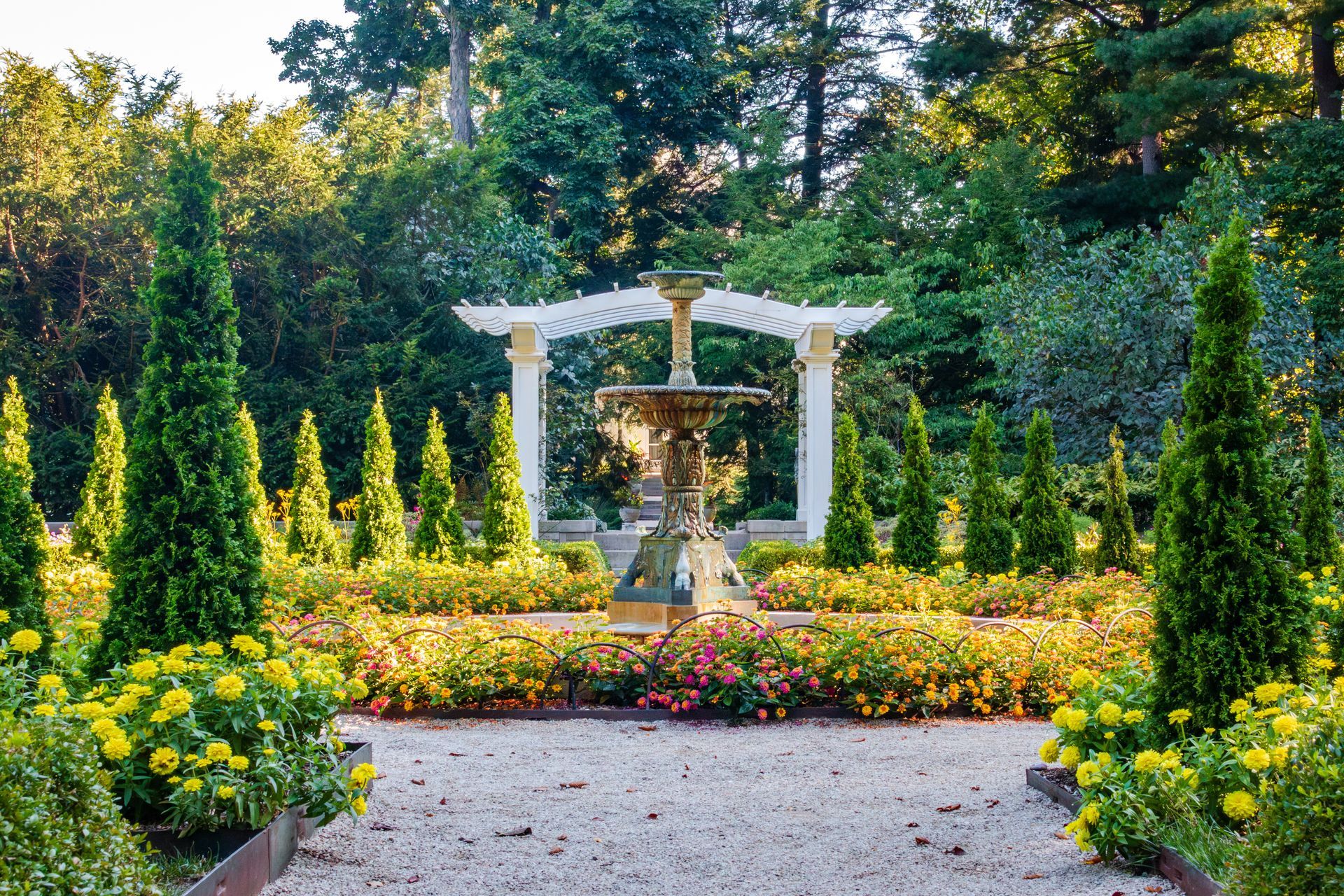 Flower garden with a white pergola and central fountain, bordered by yellow blooms and greenery.