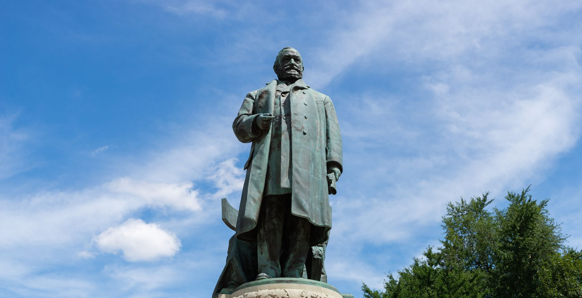 Statue of a man in a long coat standing outdoors against a blue sky and trees