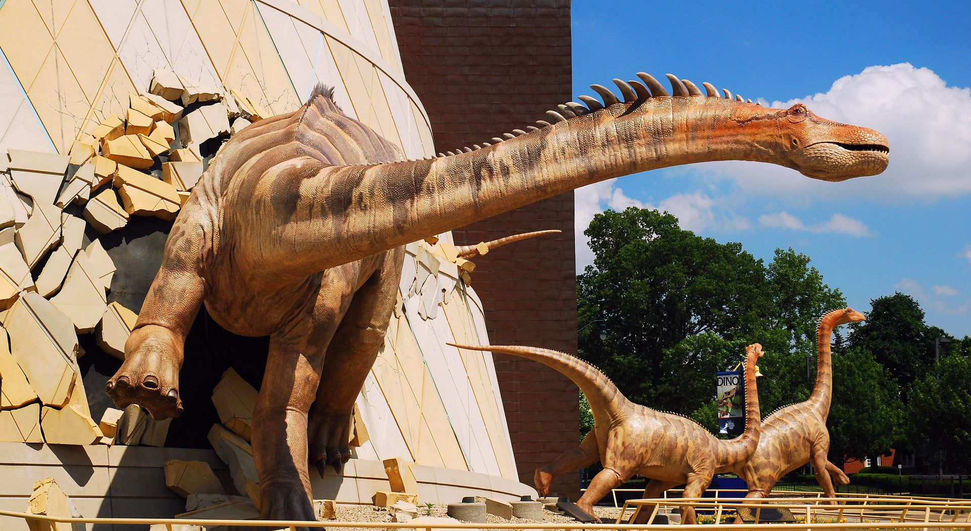 Dinosaur statues at a museum, including a large long-necked dinosaur in front of a brick building.