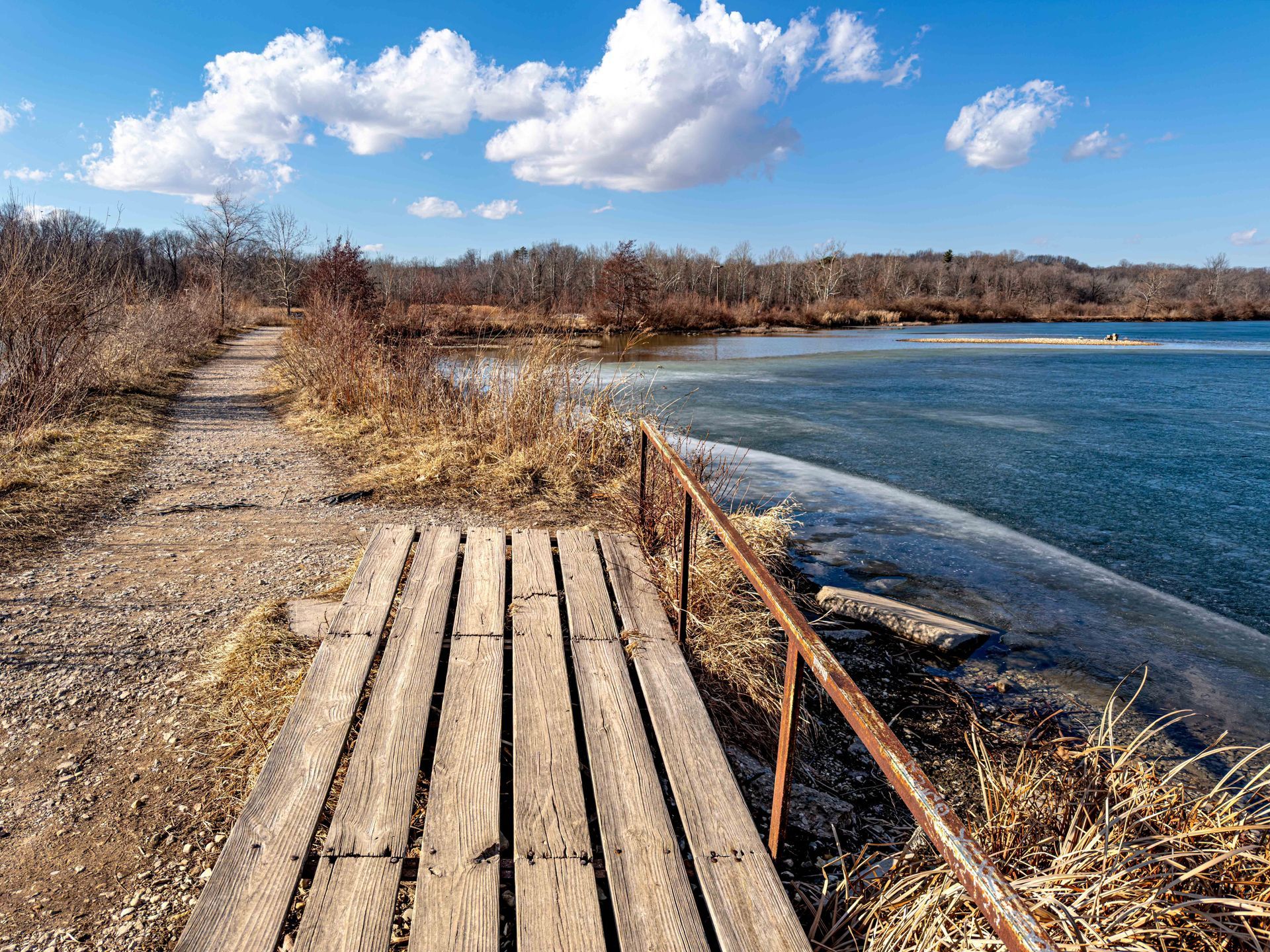 Wooden boardwalk beside a calm blue lake under a bright sky with clouds and dry brush along the shore