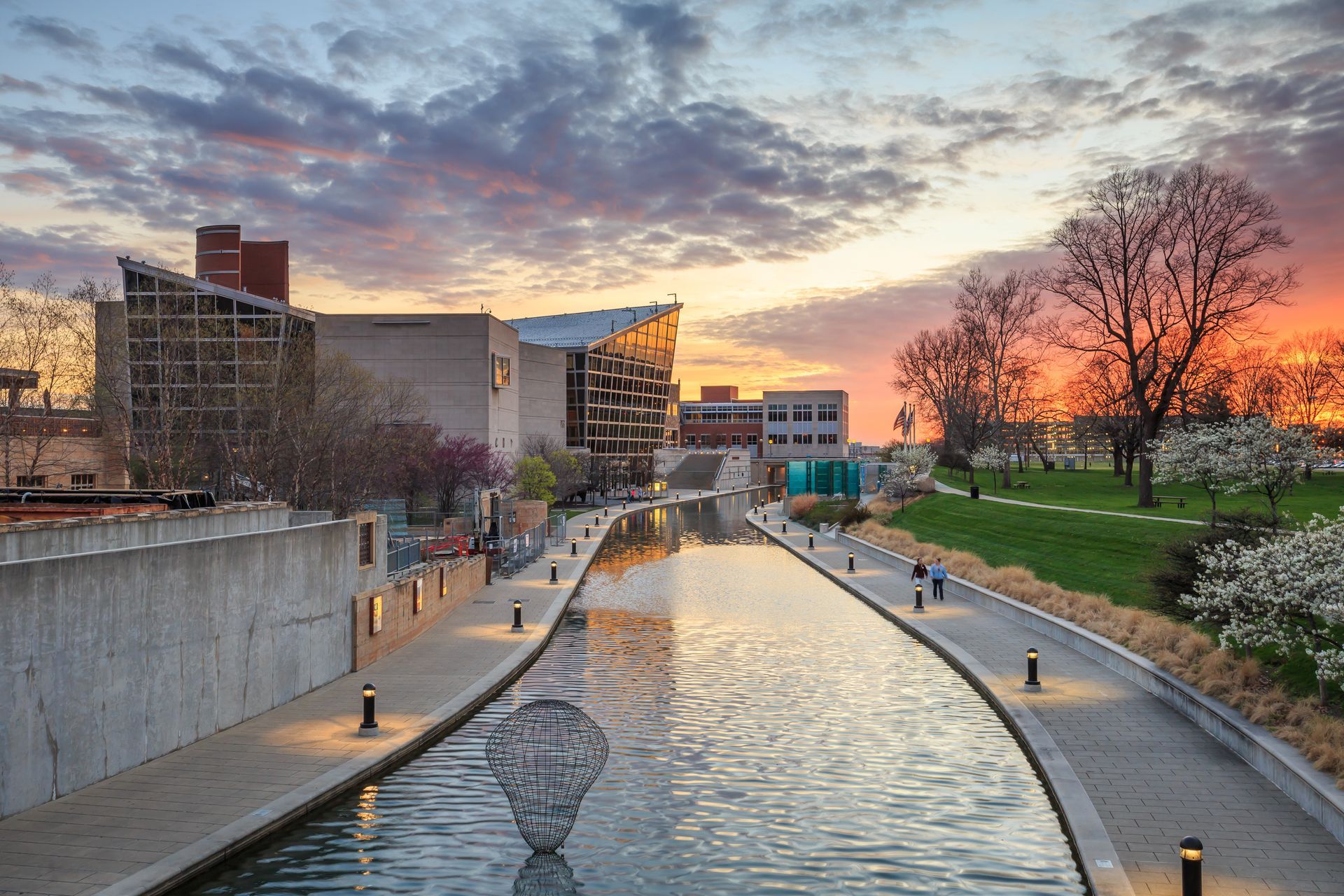 Canal at sunset with city buildings, walkway lights, and trees reflecting in calm water