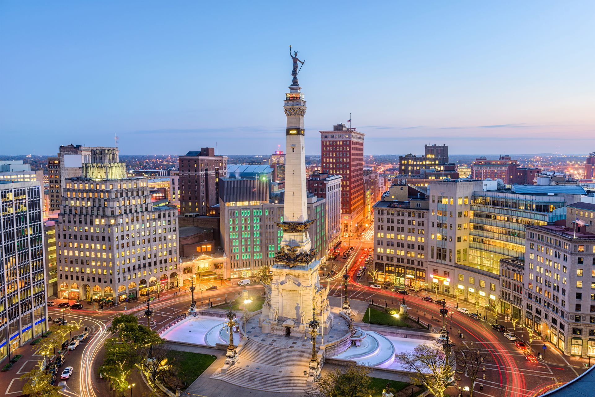 Dusk view of a city square with a central monument, lit streets, and surrounding buildings