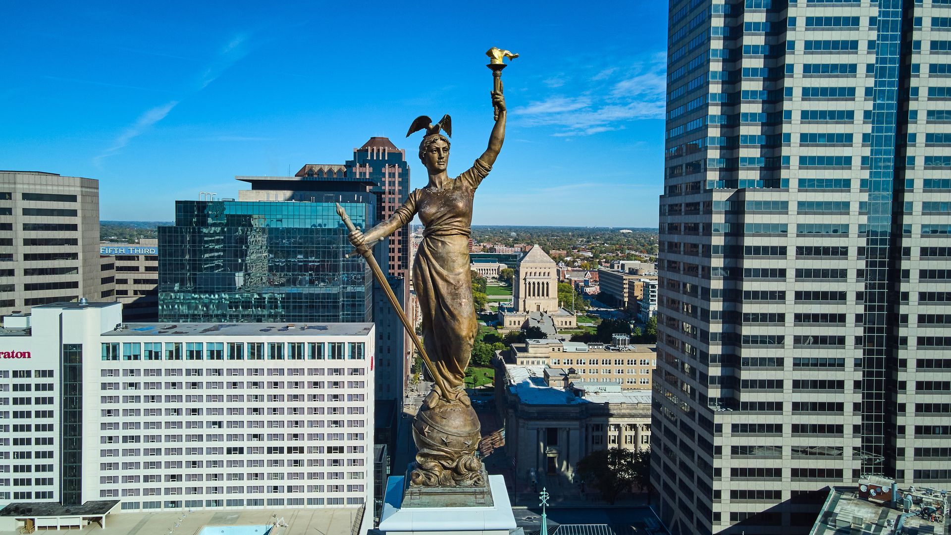 Gold statue on a rooftop in a city skyline, with tall buildings and blue sky behind it.