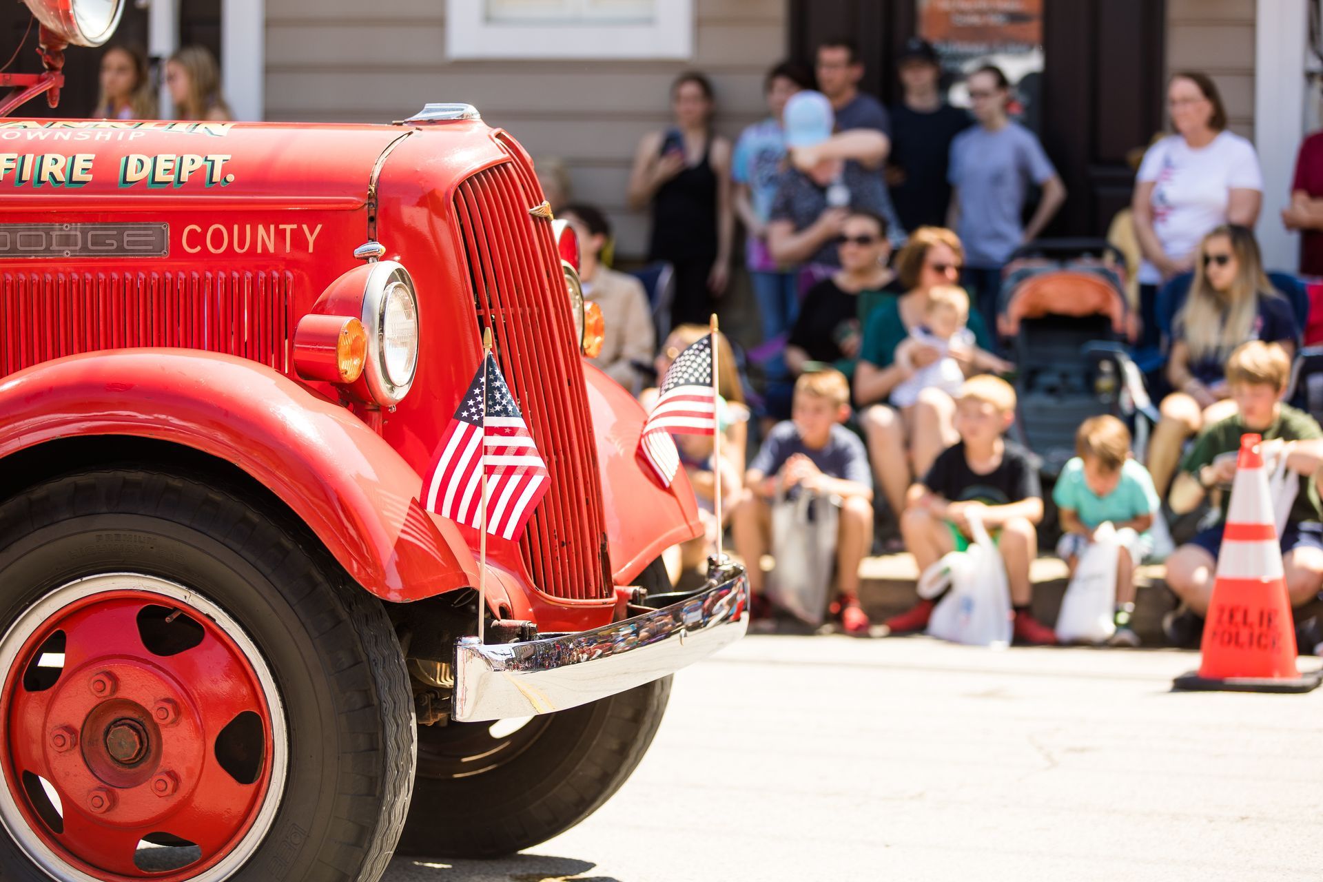 A red vintage fire truck with small American flags attached to the front grill parked during a daytime outdoor parade.