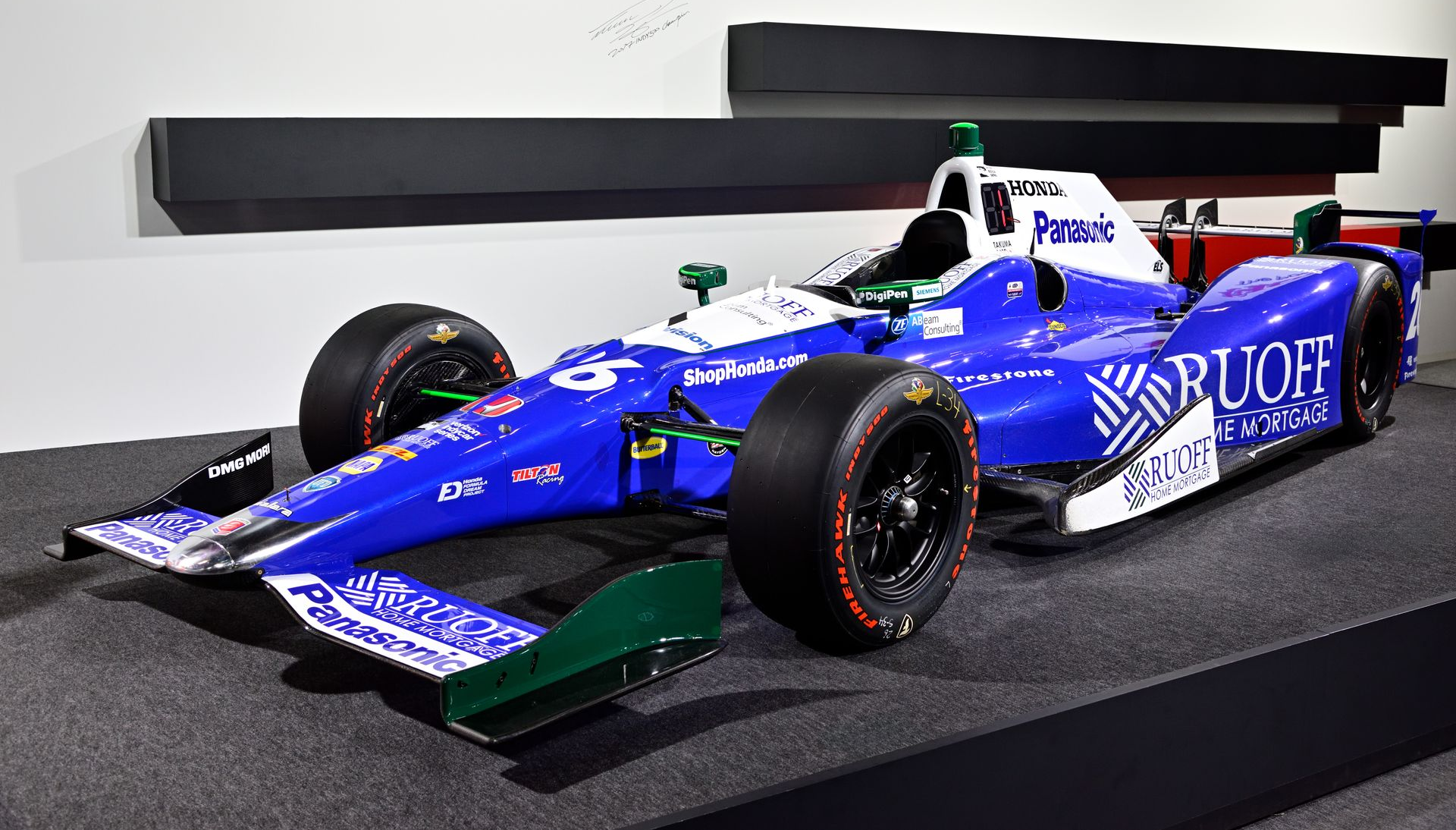 A blue and white IndyCar racer with sponsorship decals, parked on a platform in an indoor setting.