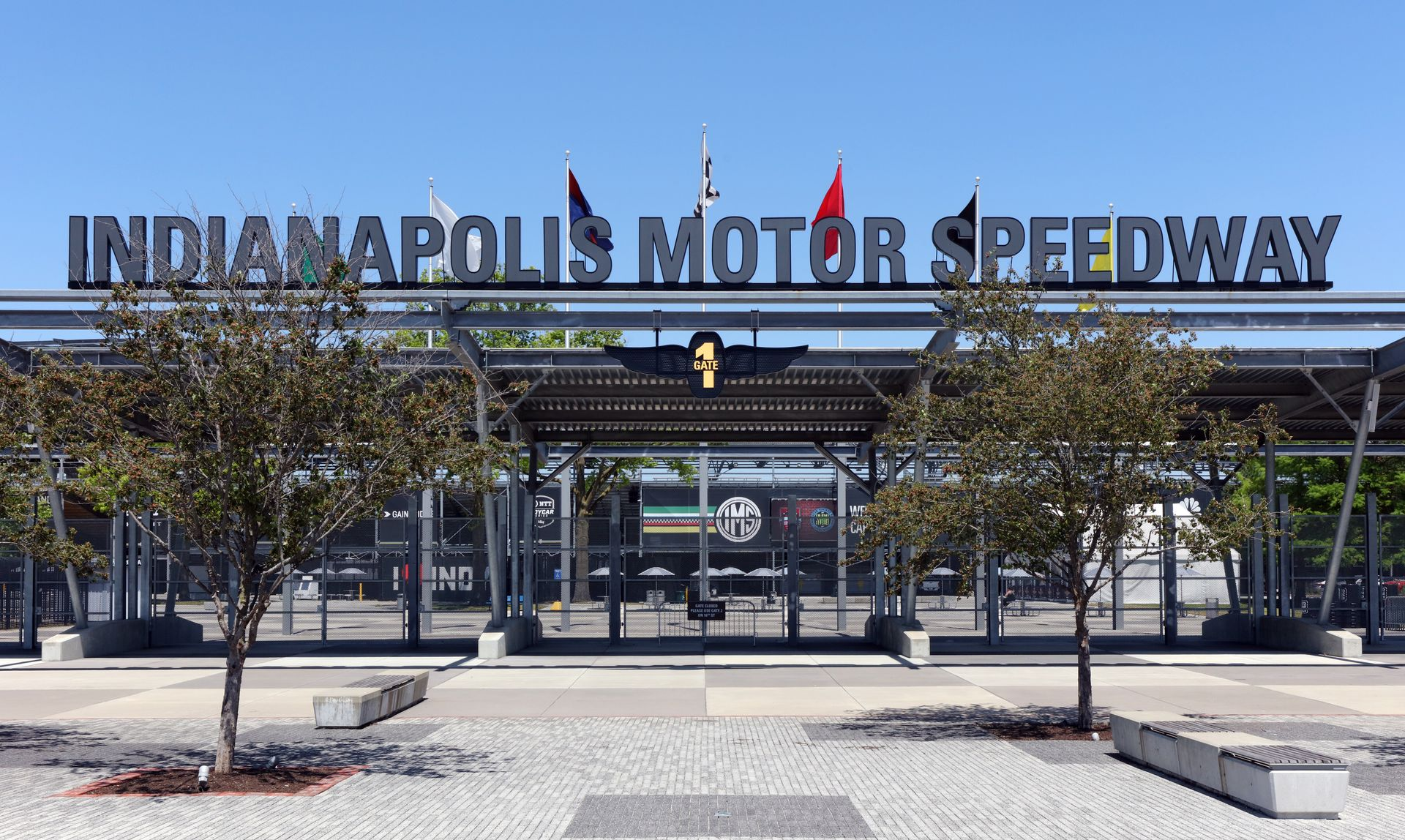 The large, iconic black metal sign for the Indianapolis Motor Speedway stands above an entrance gate under a clear sky.
