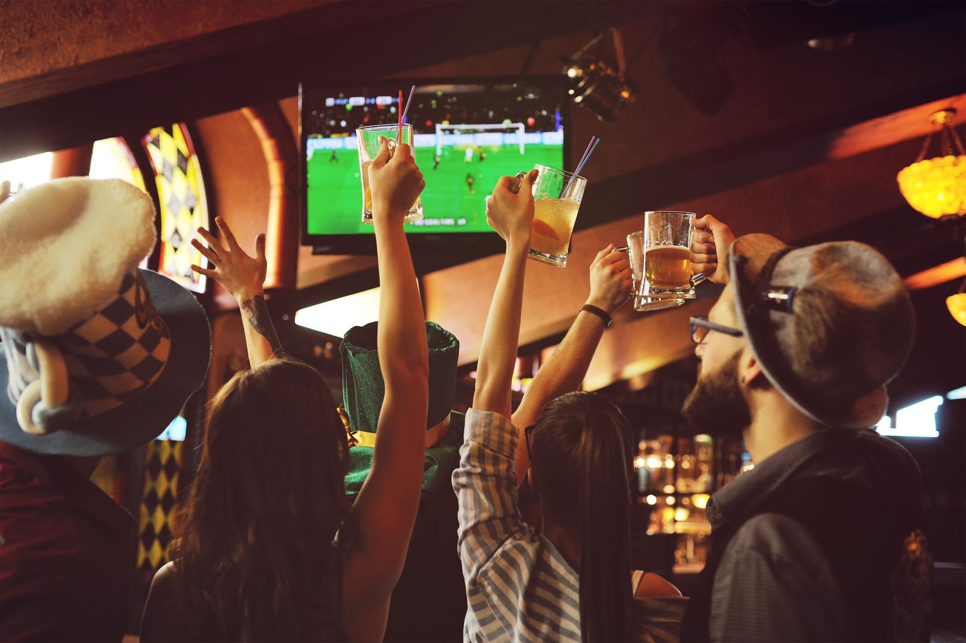 People watching a soccer match on a TV in a dim sports bar, cheering with raised hands.