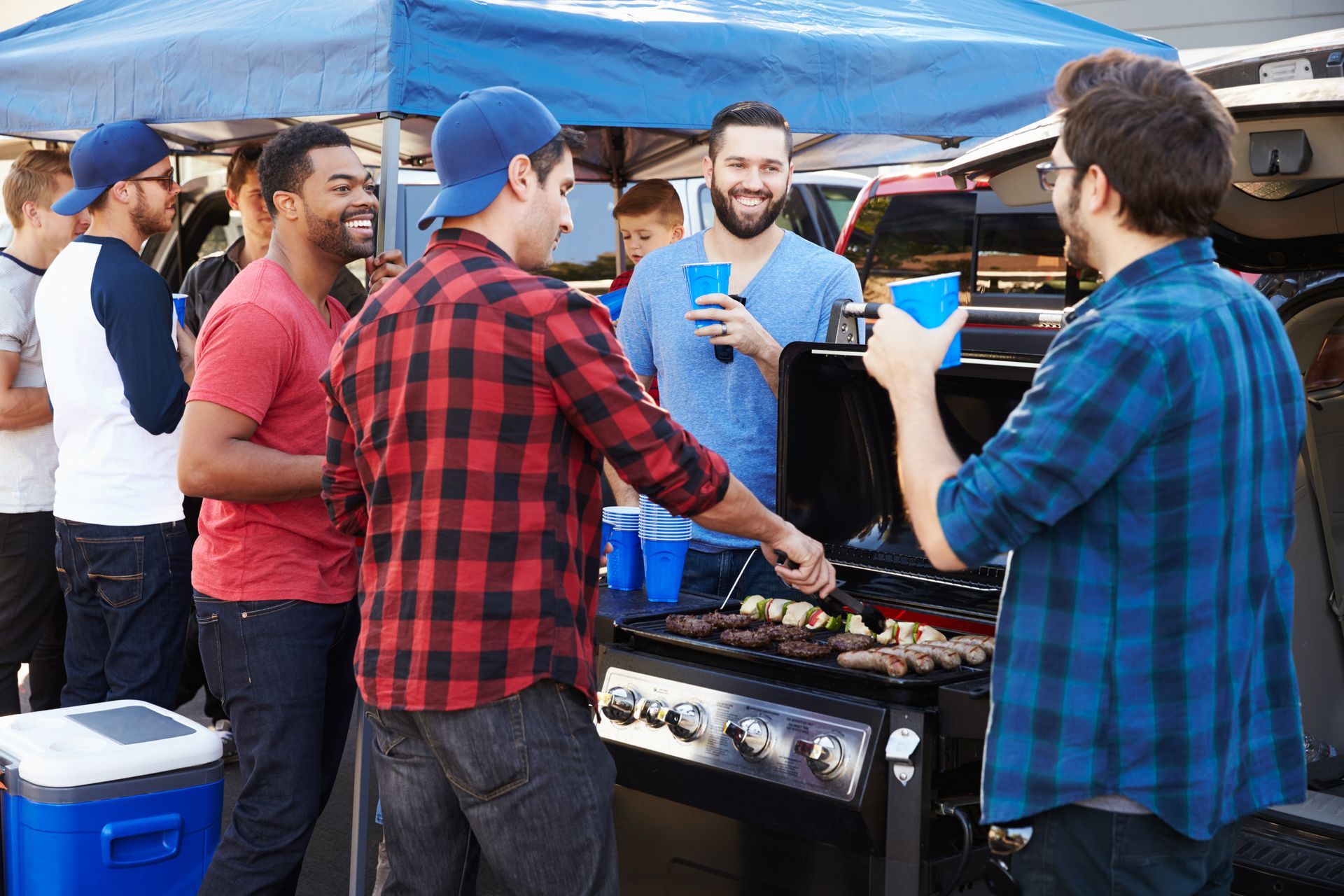 Men gathered around a grill at an outdoor cookout under blue canopies, chatting and holding drinks.