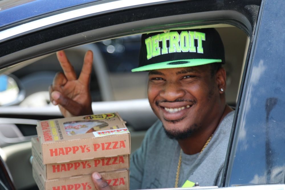A man wearing a detroit hat is holding a stack of happy pizza boxes