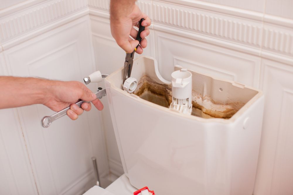 Hands repairing a broken toilet tank with exposed parts and a wrench in a bathroom.