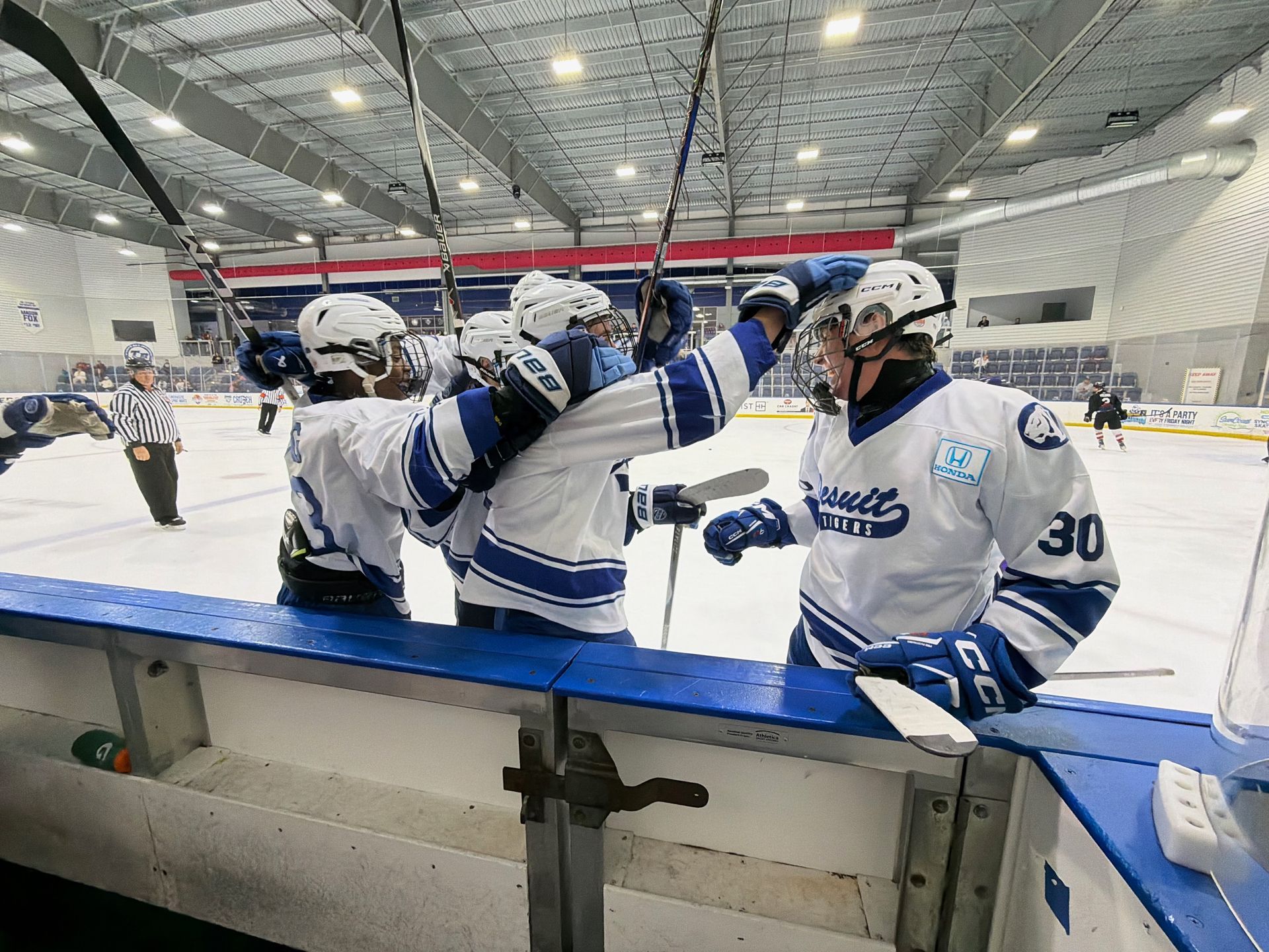 Hockey players in white and blue celebrate on the bench during an indoor ice rink game.