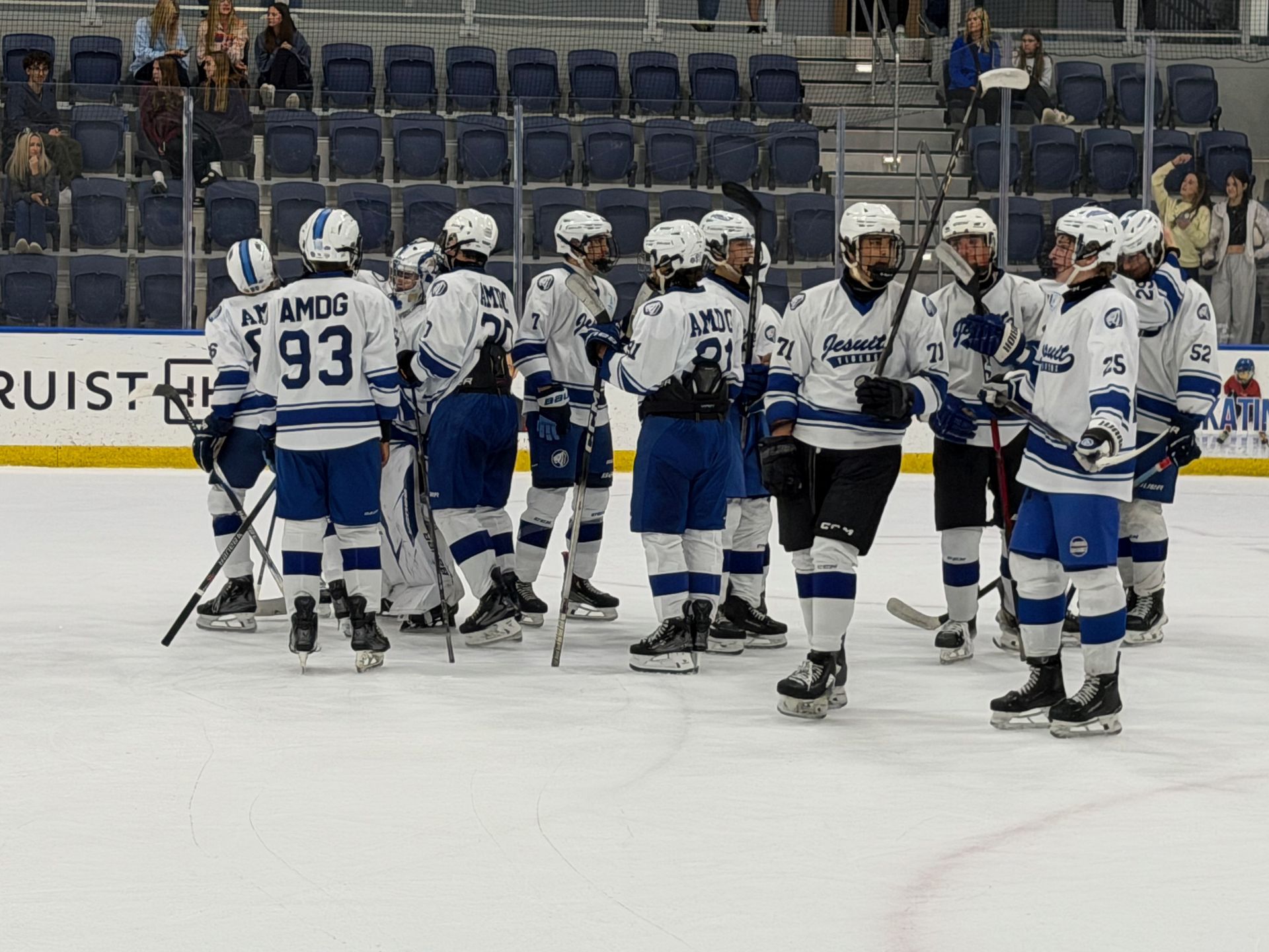 Ice hockey players in white and blue uniforms gather on the rink near the goal during a pause.