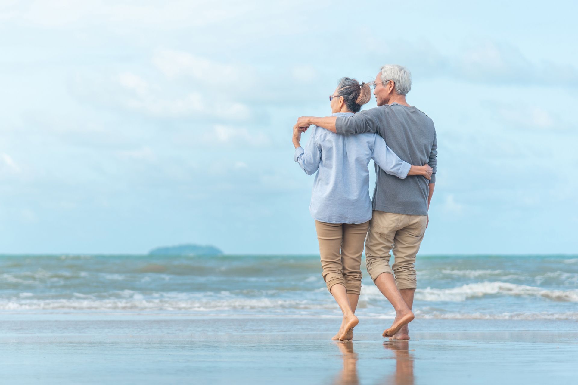 An elderly couple is standing on the beach looking at the ocean.