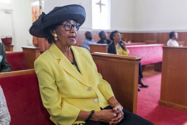 A woman in a yellow jacket and black hat is sitting in a church.
