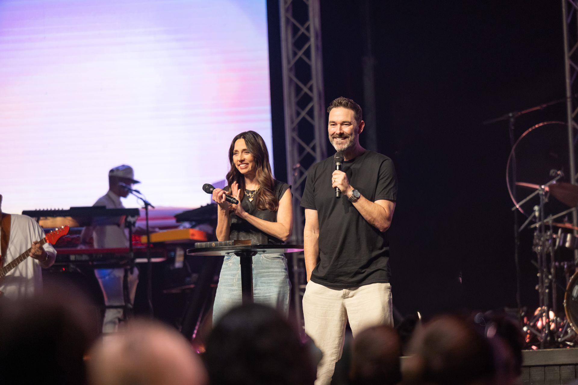 Two speakers onstage holding microphones beside a keyboard player and drum set under stage lights
