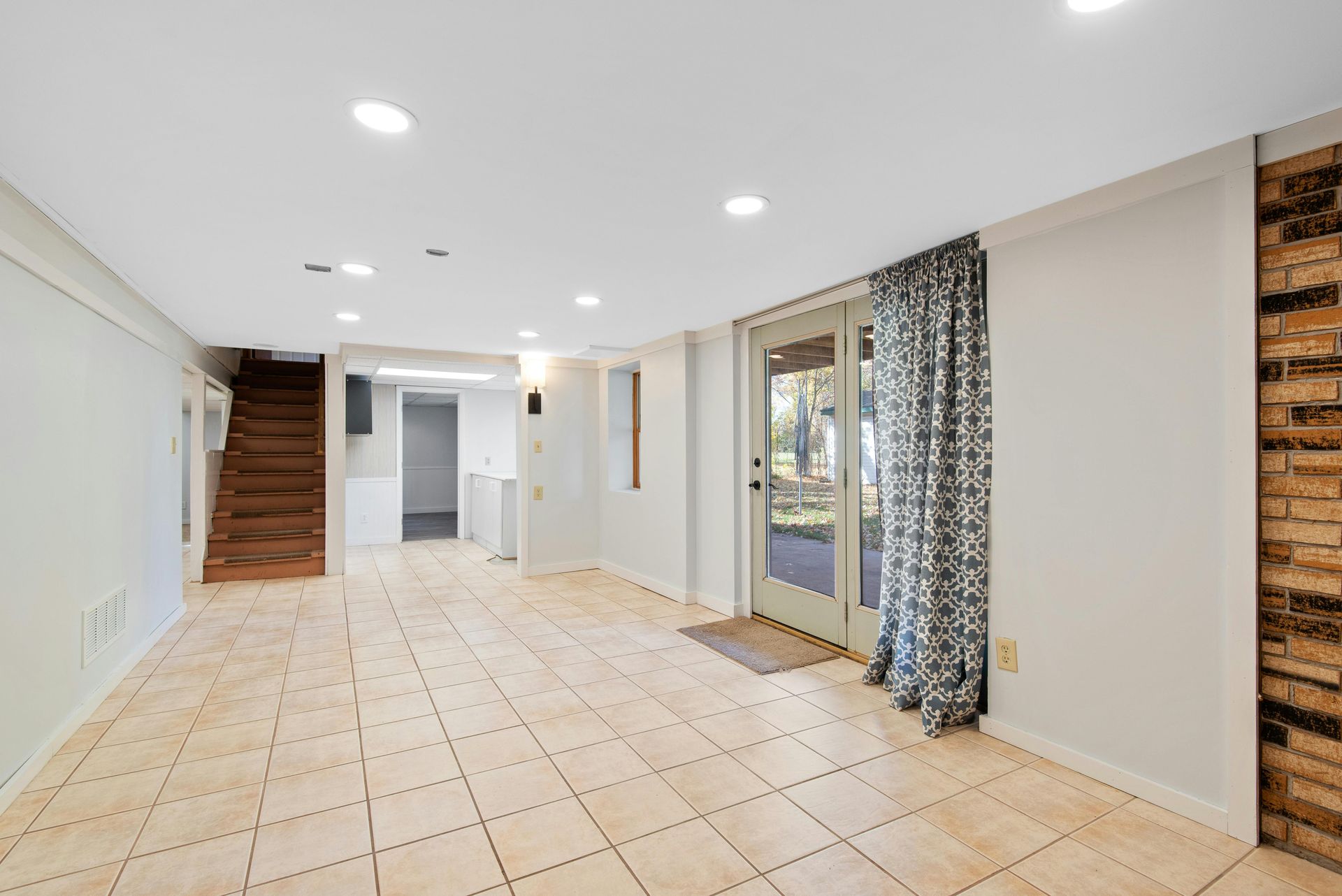 Sous-sol vide avec sol carrelé beige, murs blancs, accent en brique et une porte menant à l'extérieur.