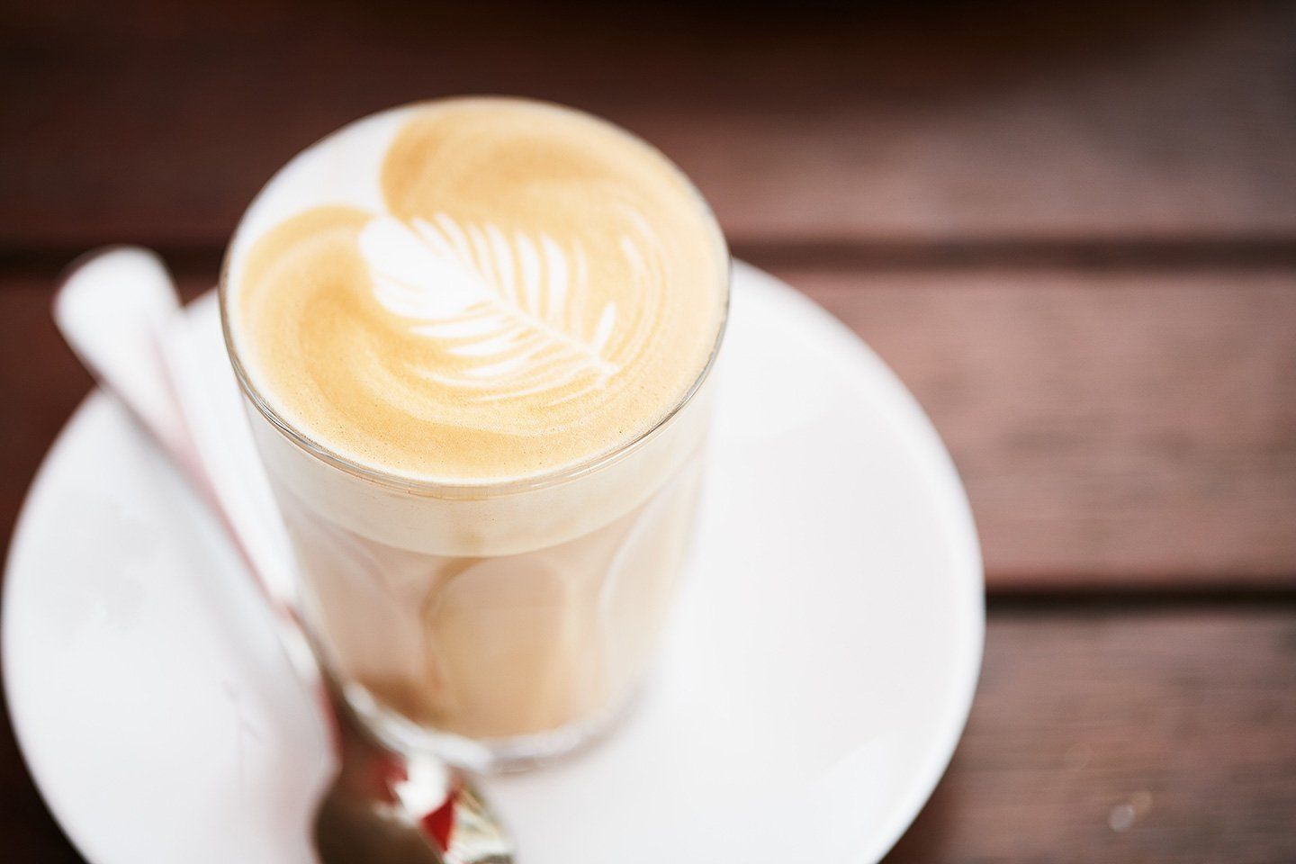 A cup of cappuccino with a spoon on a saucer on a wooden table.