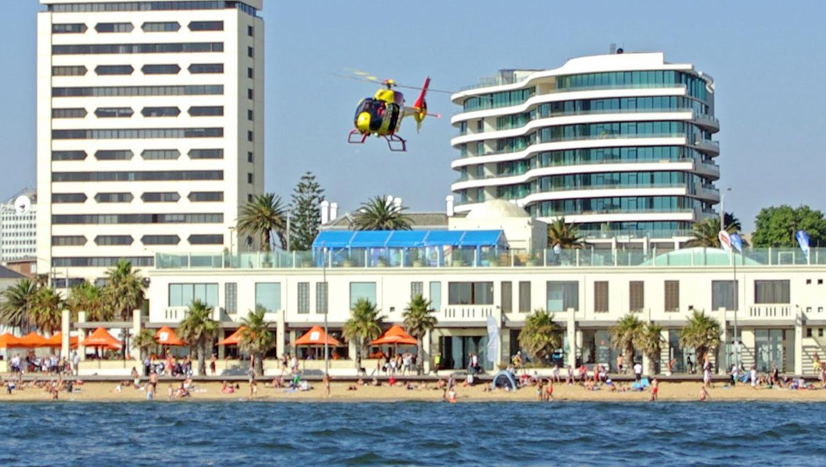 A helicopter is flying over a beach with buildings in the background
