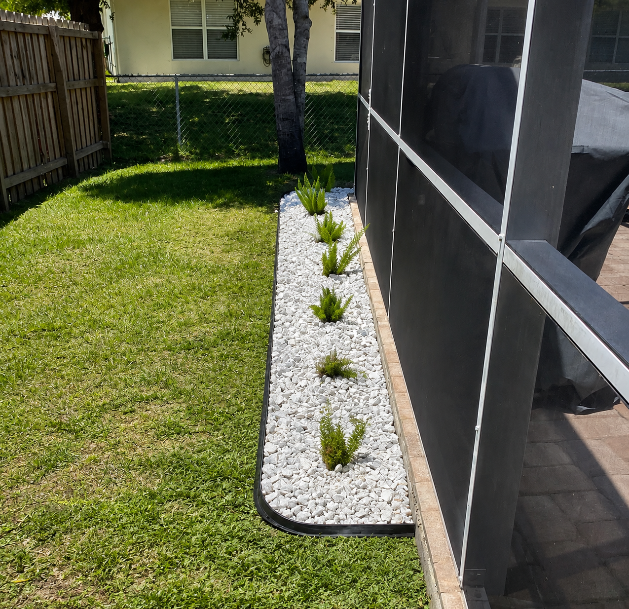 A narrow white-rock garden bed with small plants runs alongside a screened patio and green lawn.