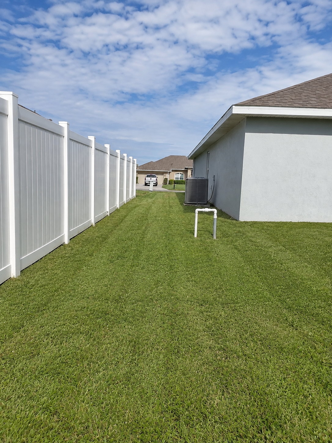 Narrow grassy side yard between a white fence and house, with a small white table near the wall.