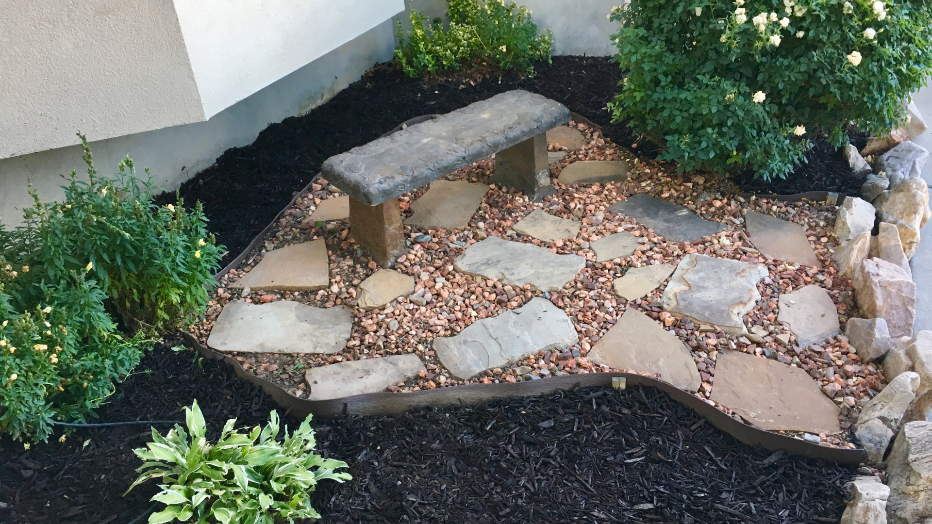 Small garden bed with stone path, bench, and green shrubs beside a house wall