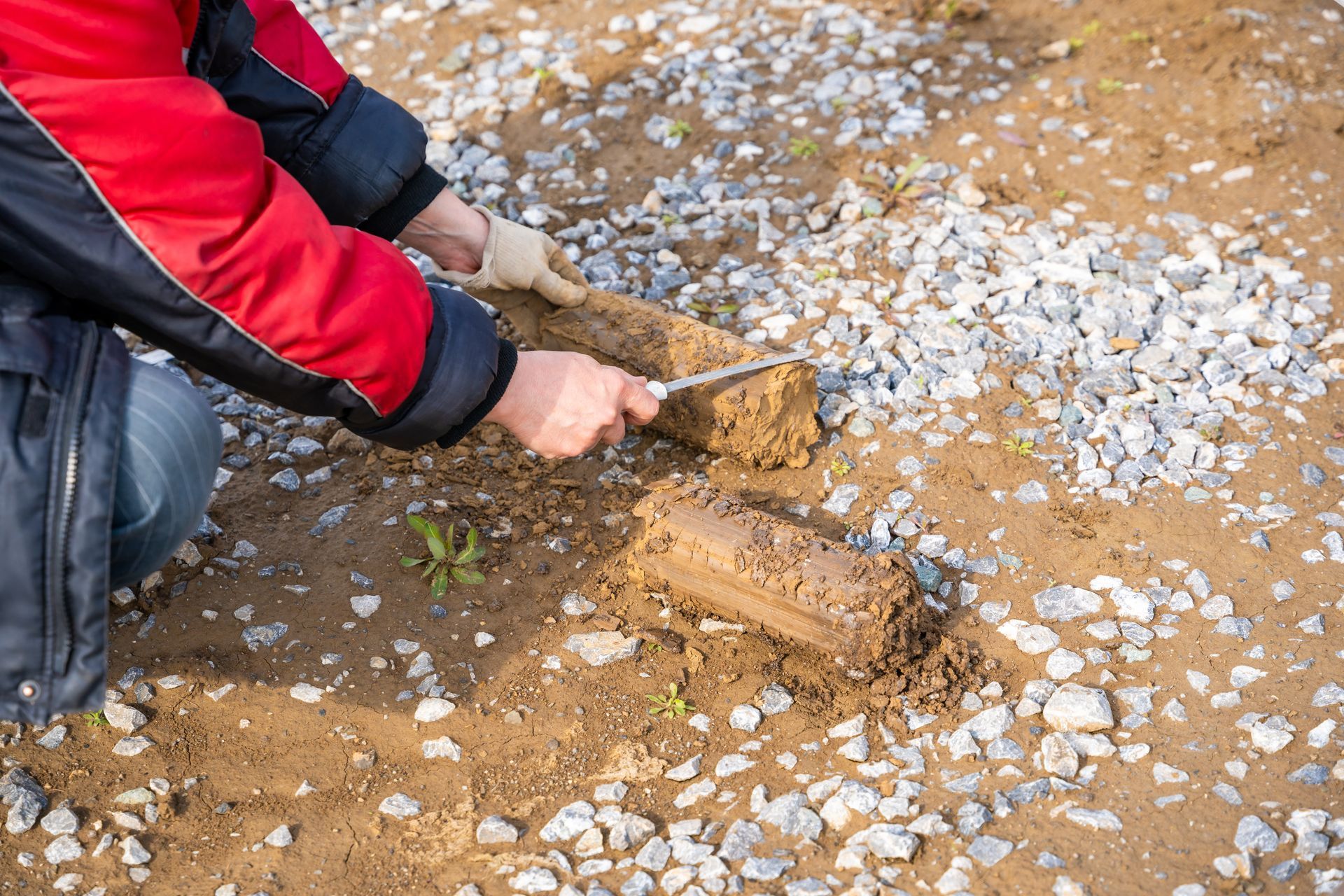 Person scraping wet mud from a shovel onto a muddy ground surface with rocks scattered around