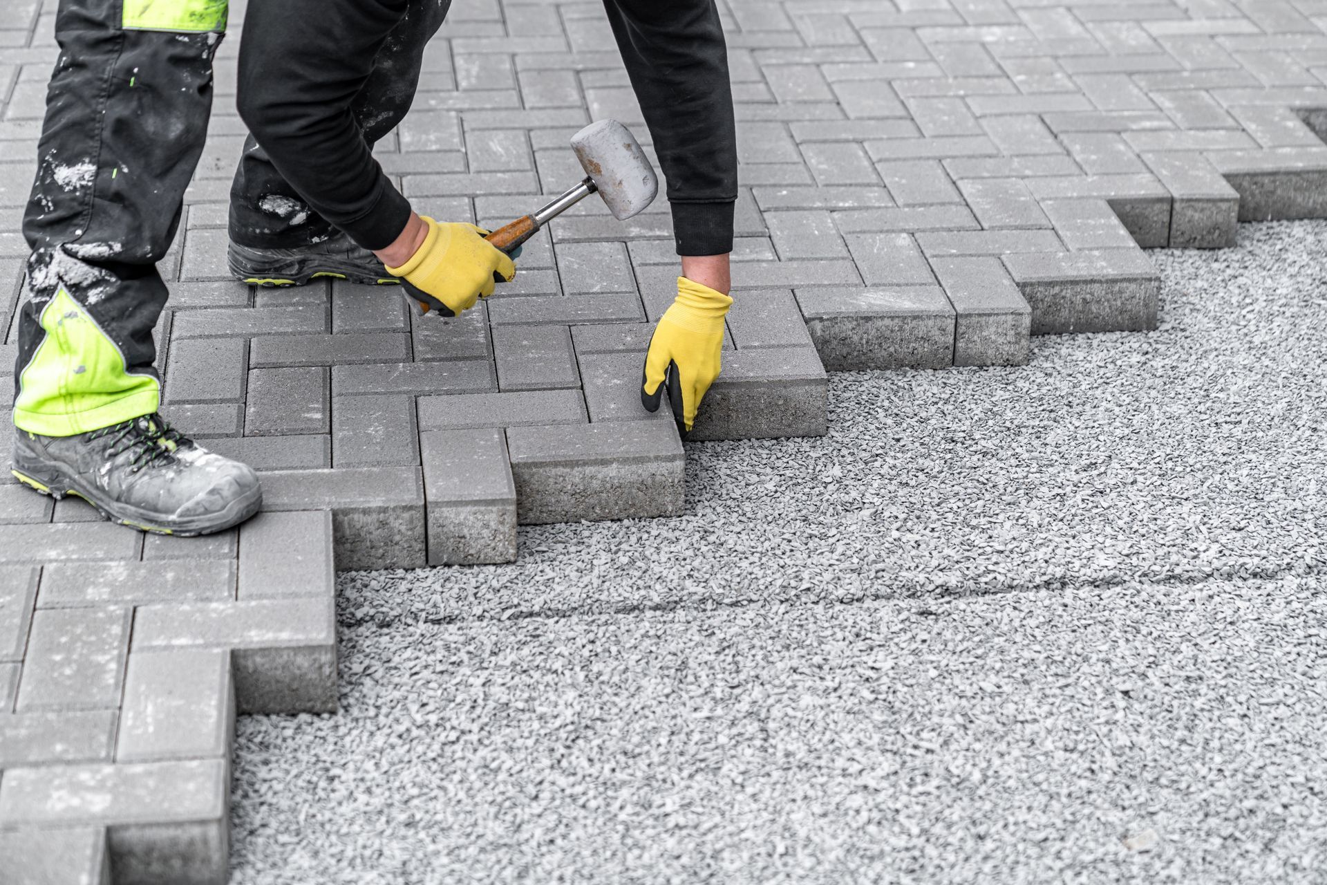 Worker laying gray pavers with a hammer and gloves on a sidewalk under construction