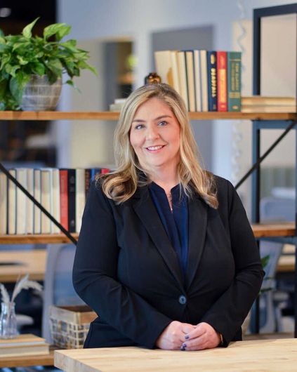 Woman standing in an office, smiling beside shelves with books and a potted plant