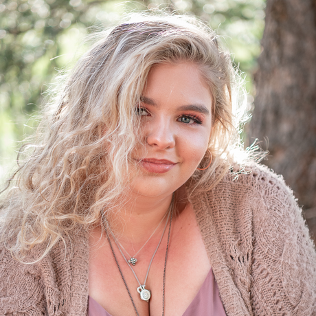 Woman with curly blonde hair in a pink top and beige cardigan, smiling outdoors near trees.