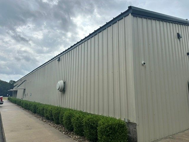 Long beige metal building with green hedges along the side under a cloudy sky