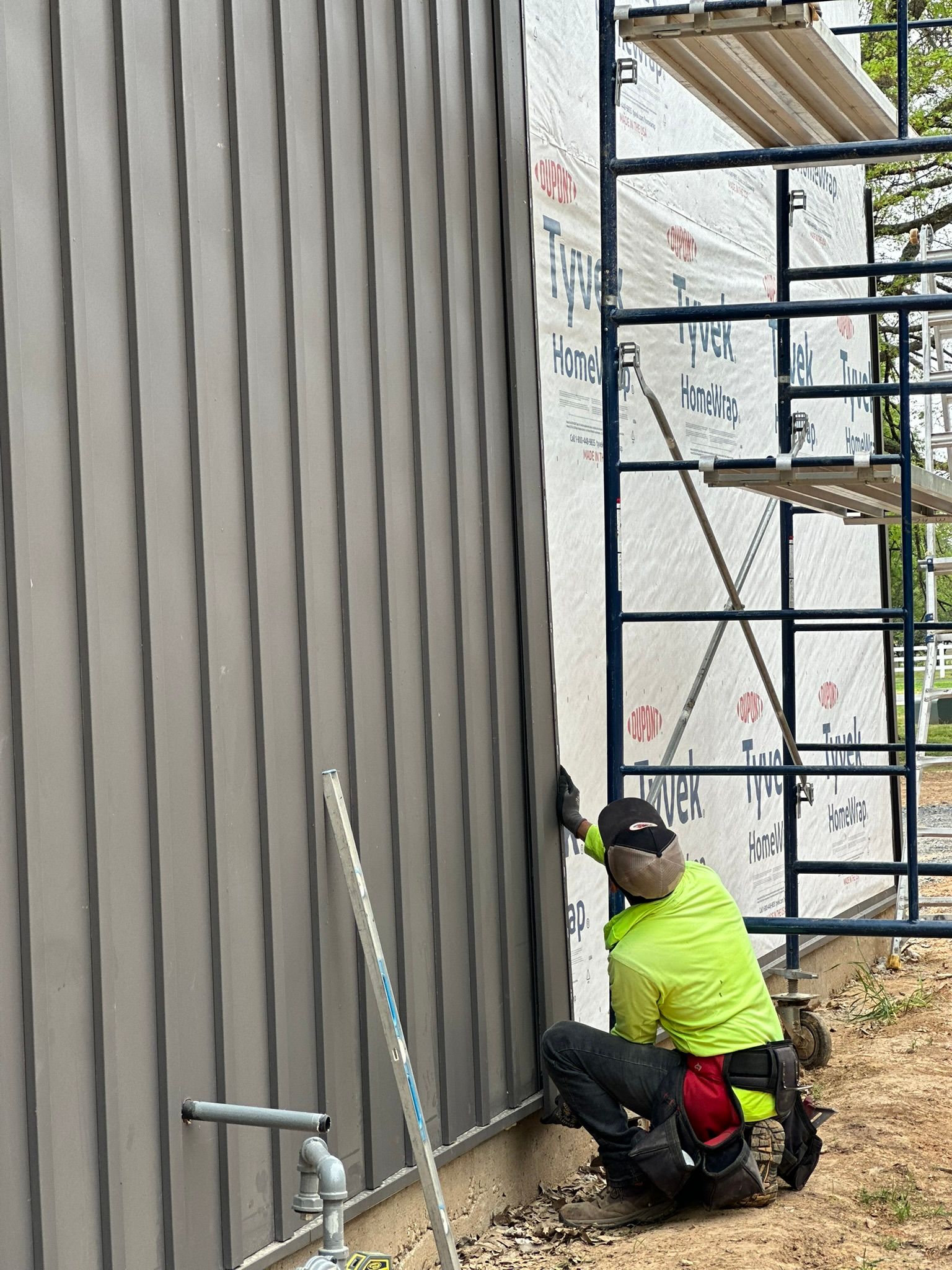 Worker in neon shirt installing gray metal siding beside scaffolding and a wall.