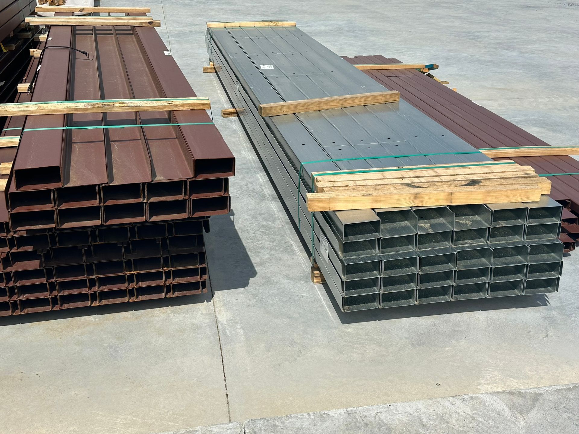Stacks of steel beams and metal panels secured with wooden braces on a concrete lot.