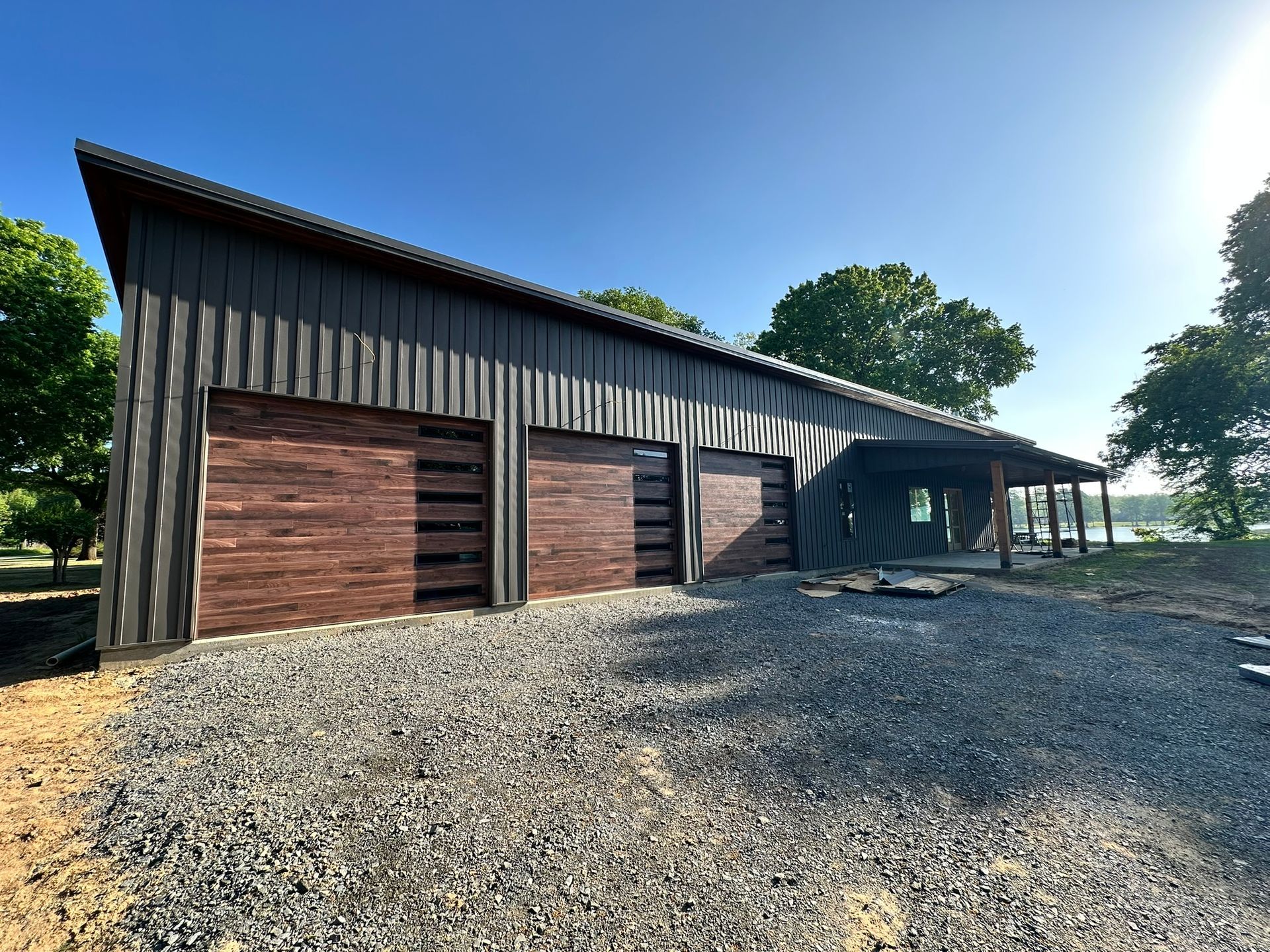 Modern rustic barn with wood siding, gravel foreground, and sunny trees in the background
