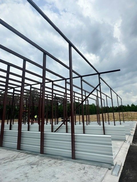 Steel frame structure under construction on a concrete slab, with corrugated metal panels and cloudy sky overhead
