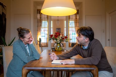 Een vrouw en een man zitten aan een houten tafel en voeren een gesprek. Notitieboekje en pen liggen naast elkaar. Warm licht. Bloemen op de achtergrond.
