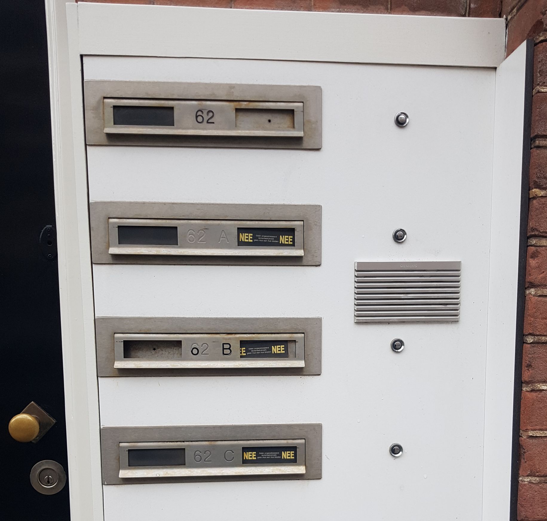 Wit paneel met vier brievenbussen, intercom en huisnummers 62 en 6. Gemonteerd op een bakstenen muur.
