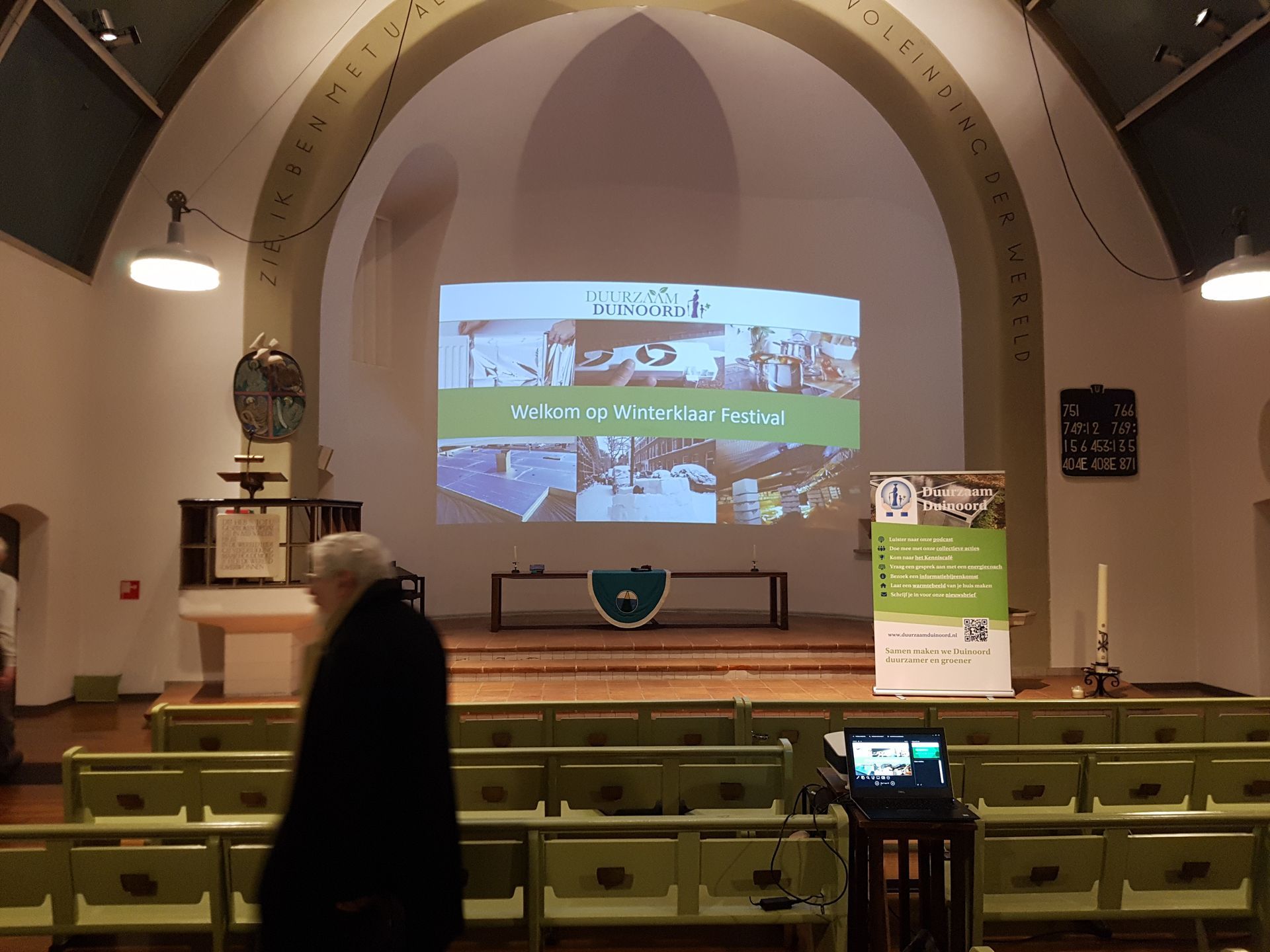 Een persoon loopt een kamer binnen met een projectiescherm. Er staat een podium vooraan met een spandoek en een presentatie.