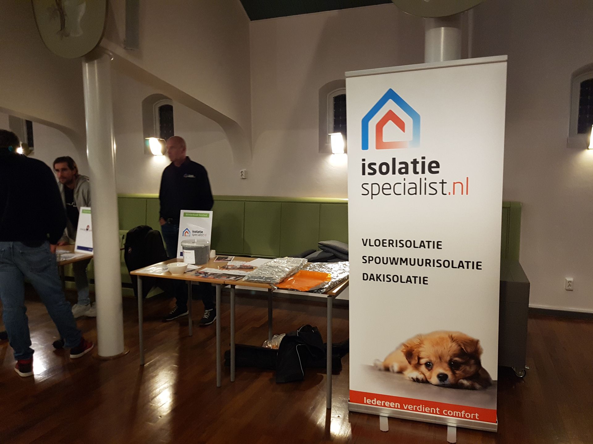 Een stand op een beurs voor bouwisolatie; een banner met logo en een puppy. Mannen staan ​​bij een tafel met brochures.