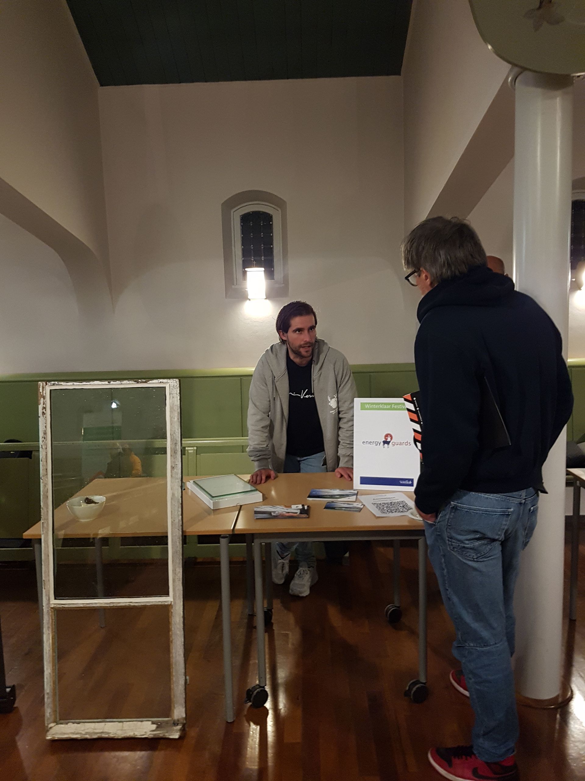 Een man zit aan een tafel met een andere man en bekijkt een vitrine; links is een oud raamkozijn te zien. De kamer heeft een houten vloer.