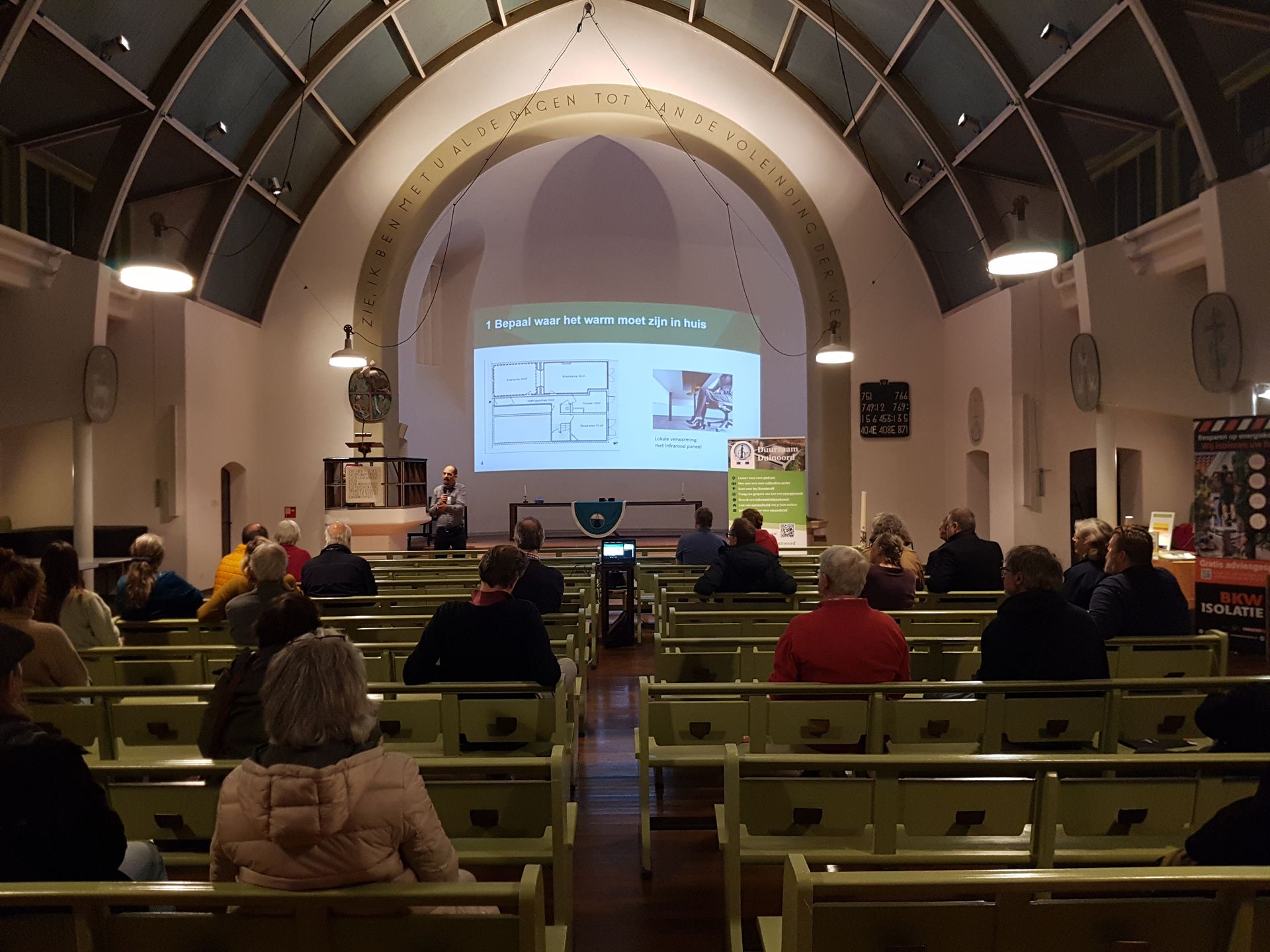 Presentatie in een kerk. Het publiek zit en kijkt naar een diavoorstelling op een scherm.