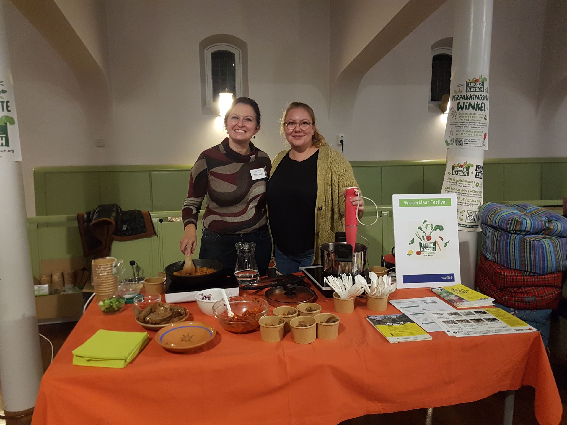 Twee vrouwen bij een eetkraam. Op een oranje tafel liggen proefhapjes en brochures. Binnen in een zaal hangen posters aan de muur.