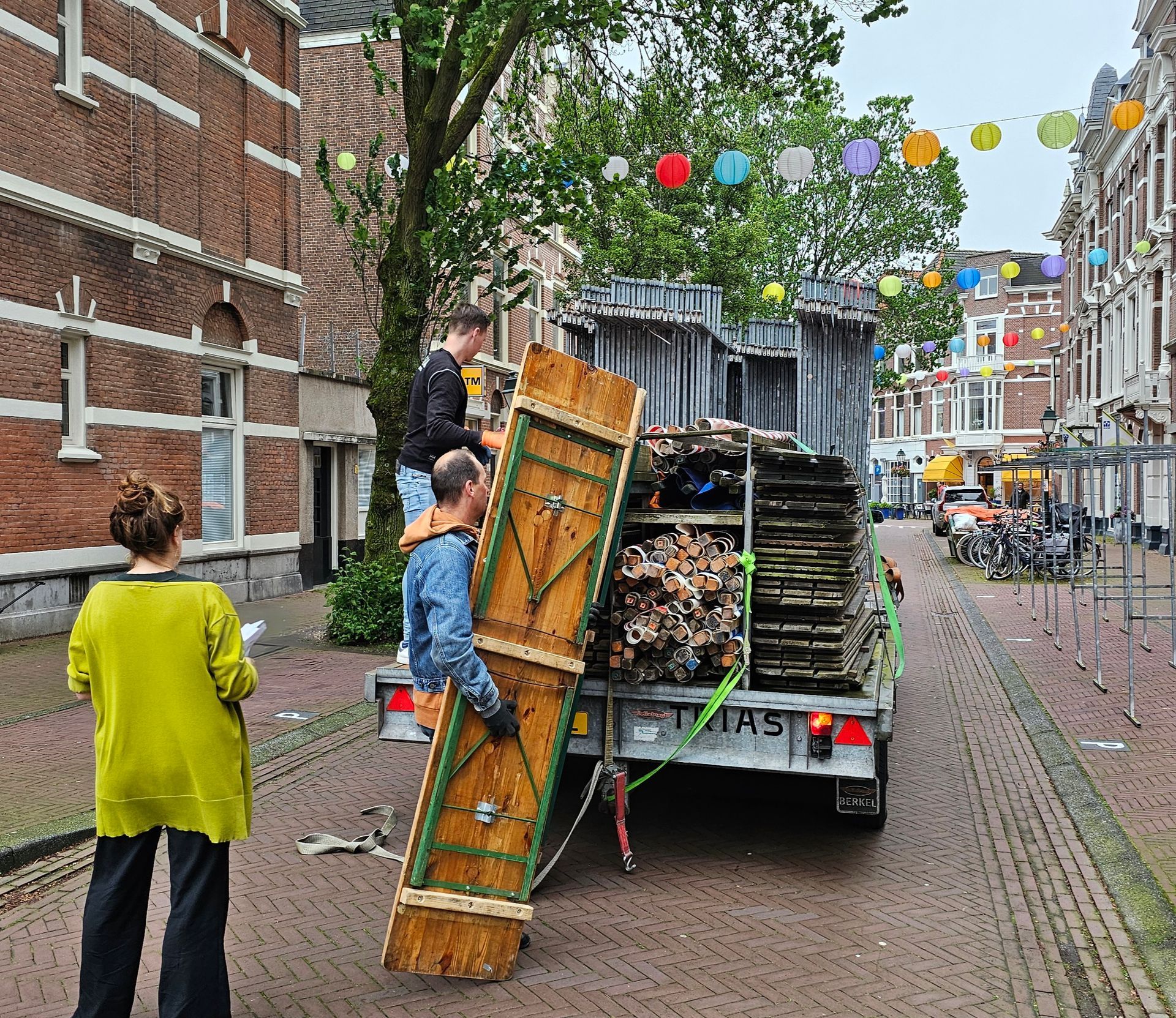 Arbeiders lossen houten doodskisten uit een vrachtwagen op een straat met kinderkopjes, versierd met kleurrijke vlaggen.