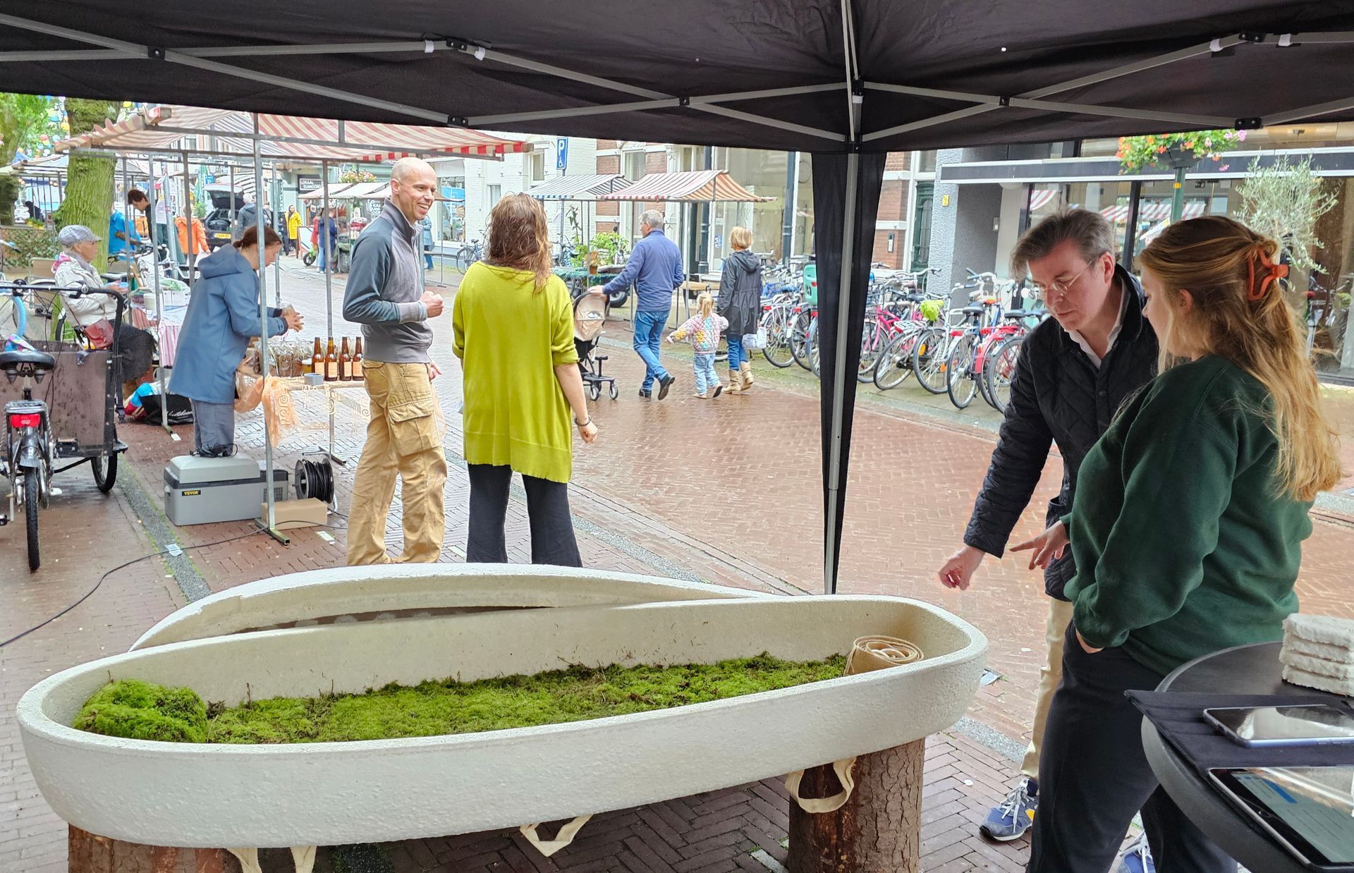 Mensen staan ​​onder een tent naast een grote plantenbak op een openluchtmarkt, met fietsen op de achtergrond.