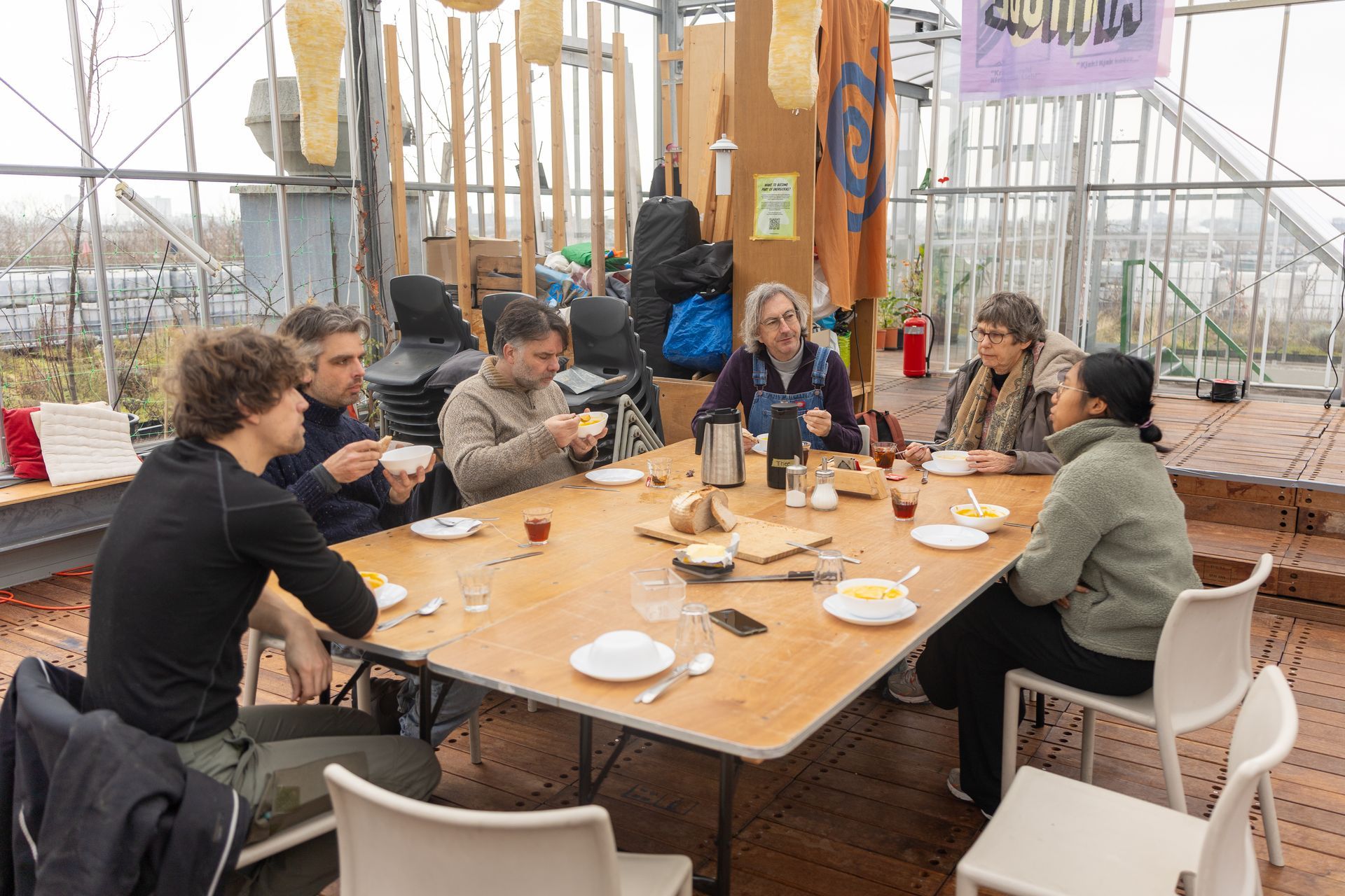 Mensen zitten rond een houten tafel in een kas, te eten en te praten met kopjes en borden.