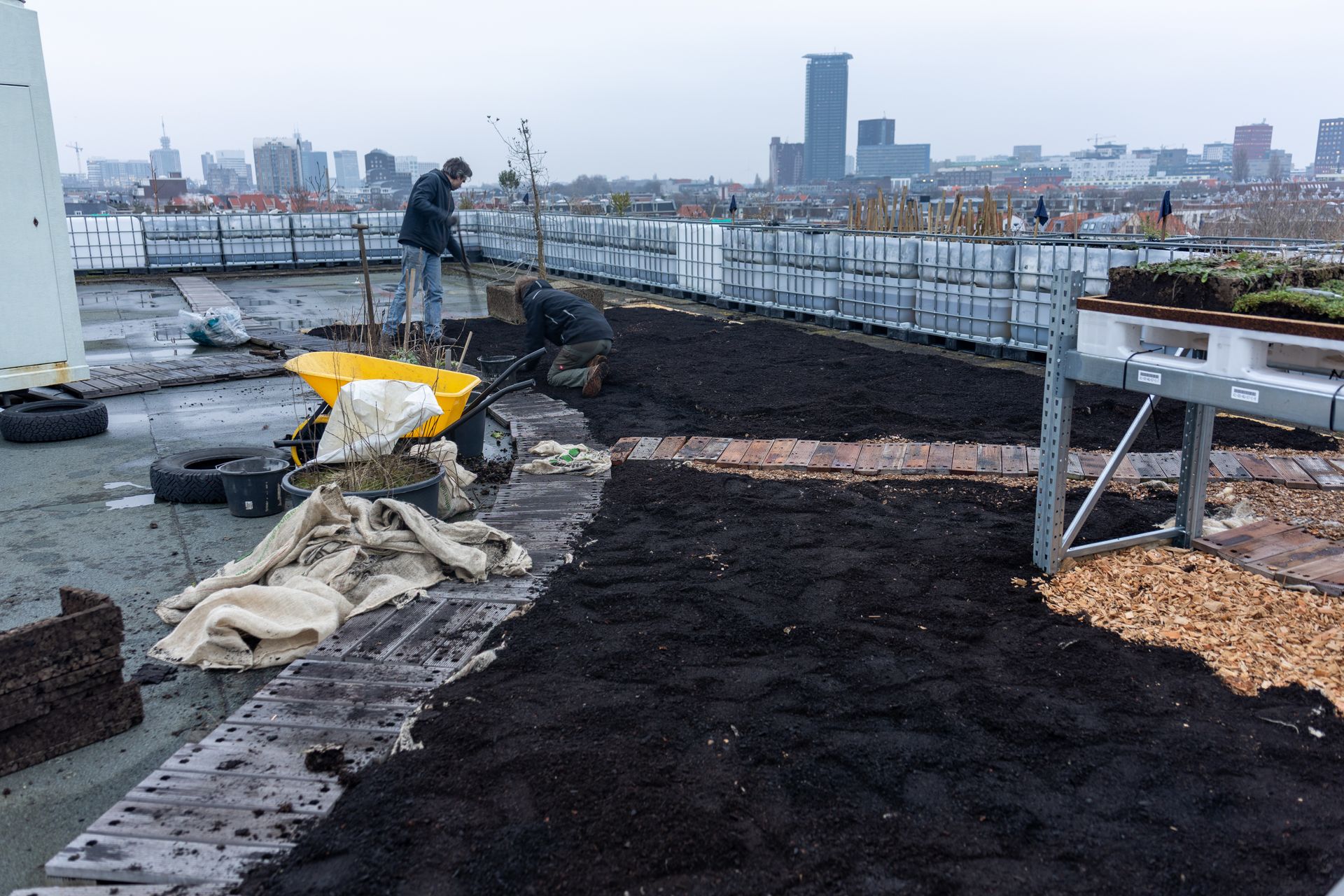 Daktuin in aanbouw met donkere grondbedden, zeilen, gereedschap en de skyline van de stad op de achtergrond.