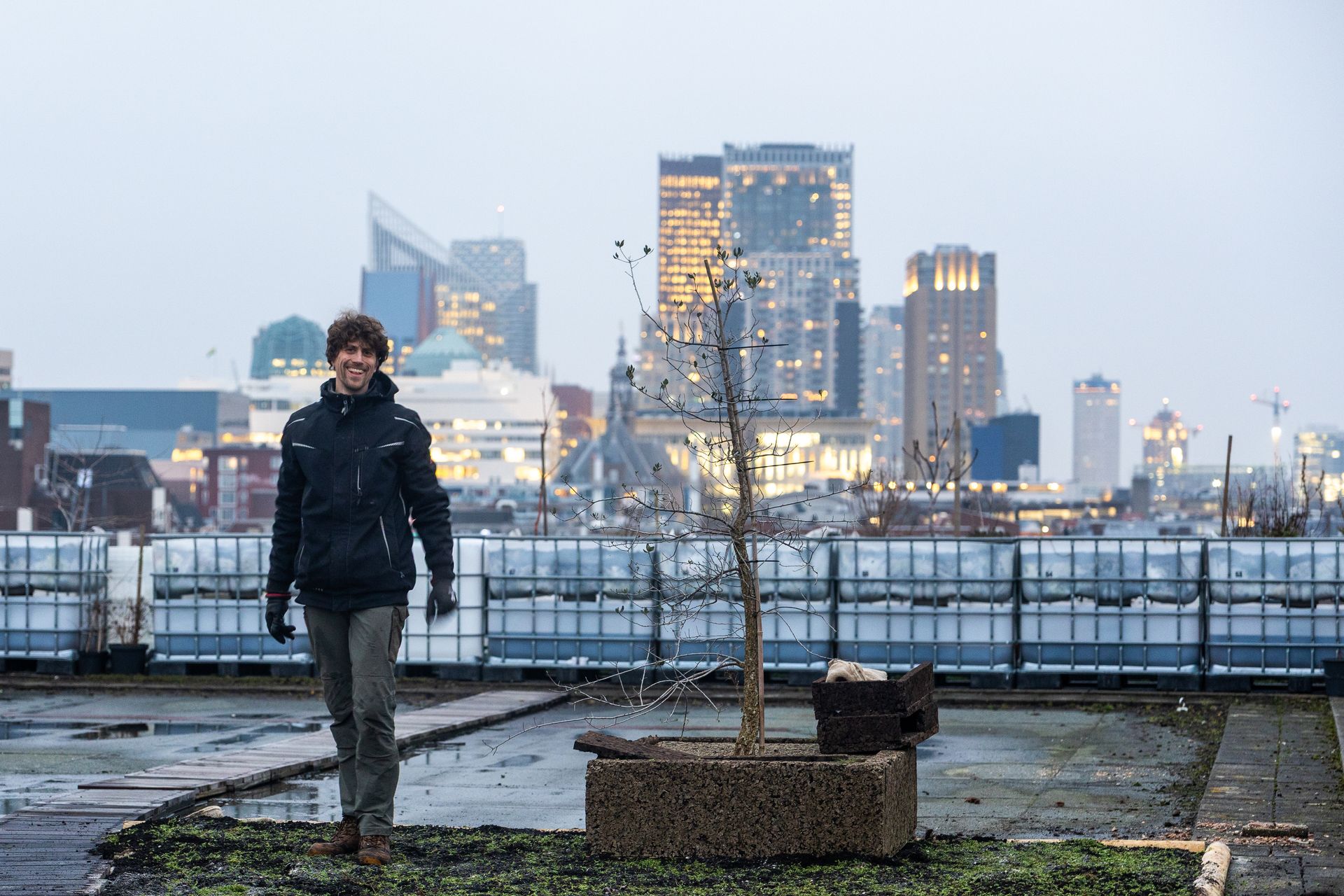 Een persoon loopt over een dakterras met op de achtergrond de skyline van de stad bij schemering.